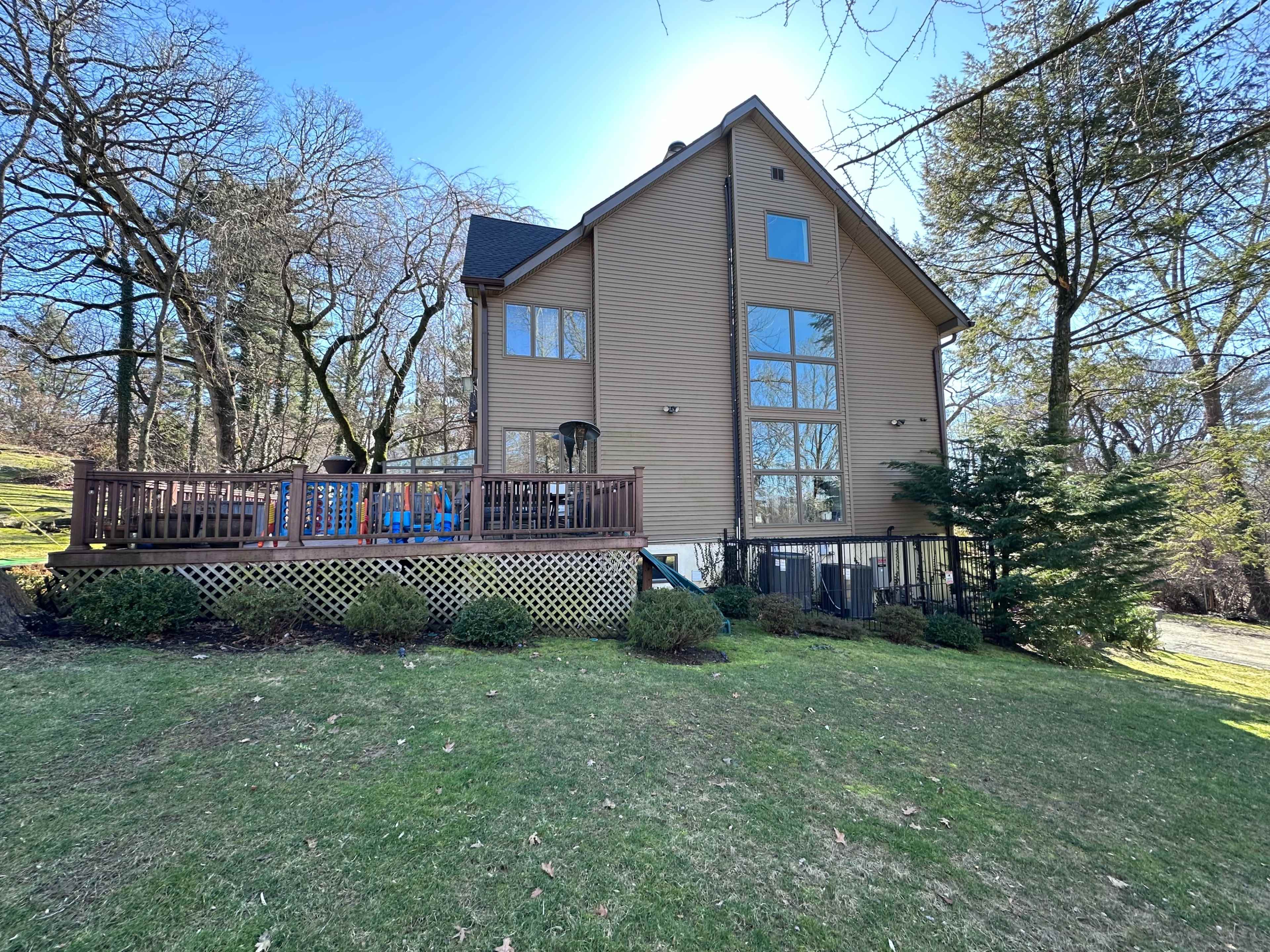 A large two-story house with a wooden deck and multiple windows is situated on a grassy lawn surrounded by trees.