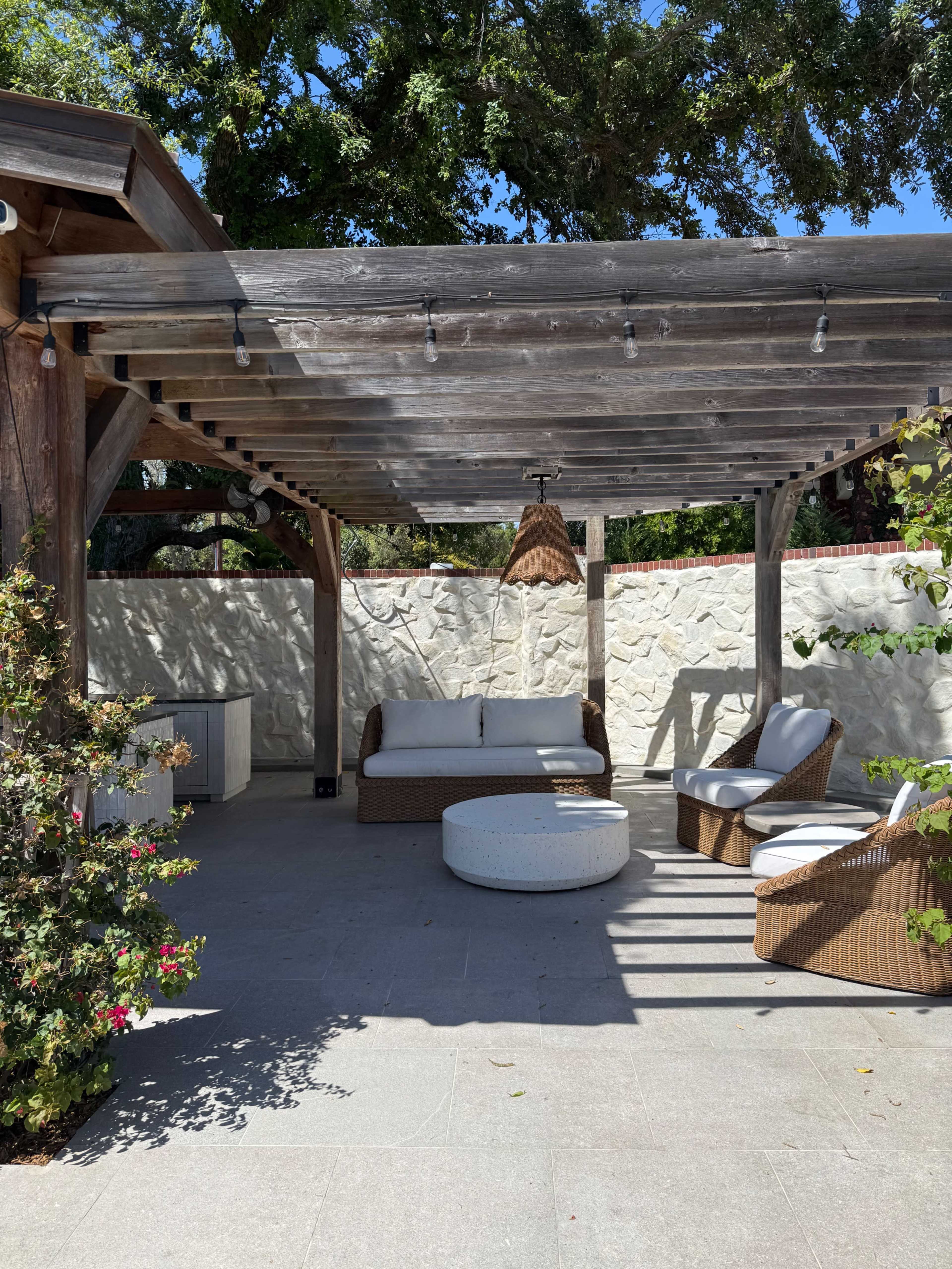 A shaded outdoor seating area features a circular table, two armchairs, and a couch beneath a wooden pergola.