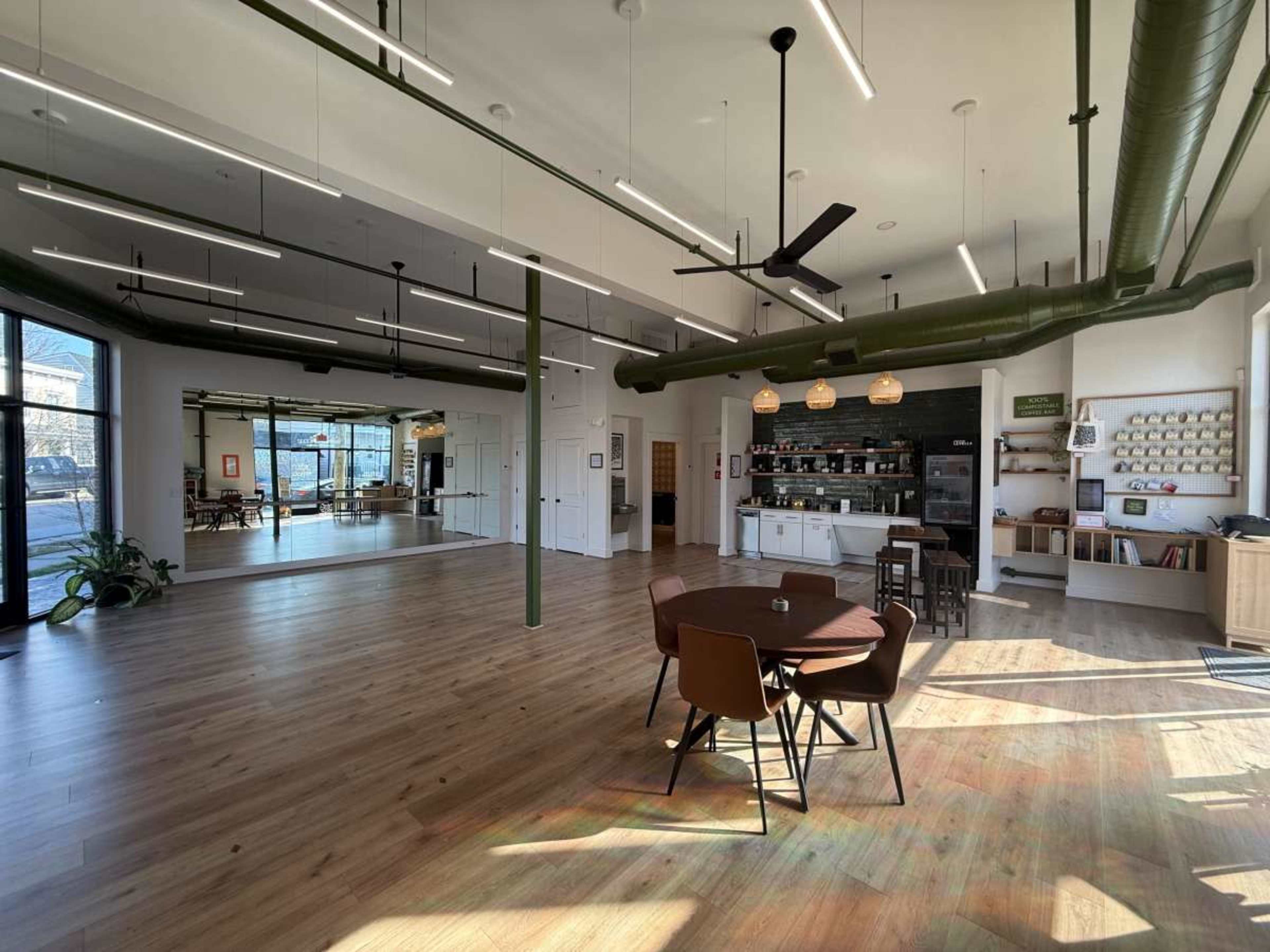 The image shows a spacious, well-lit room with wooden flooring, featuring a seating area with a table and chairs, a small kitchen area, and a large mirrored wall reflecting the space.