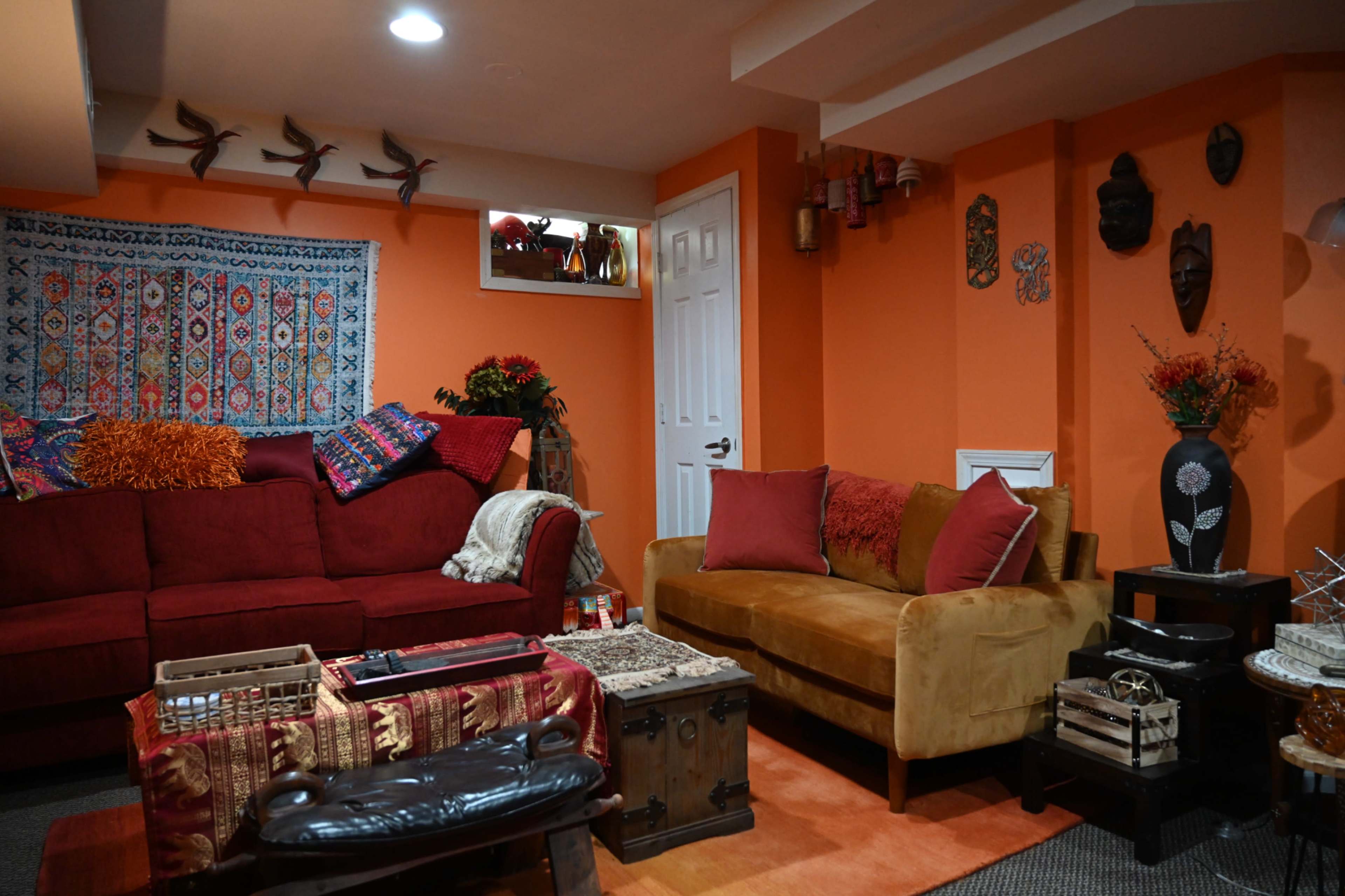 The image shows a cozy living room with orange walls, a red couch, a mustard-yellow sofa, and various decorative items on the walls and shelves.