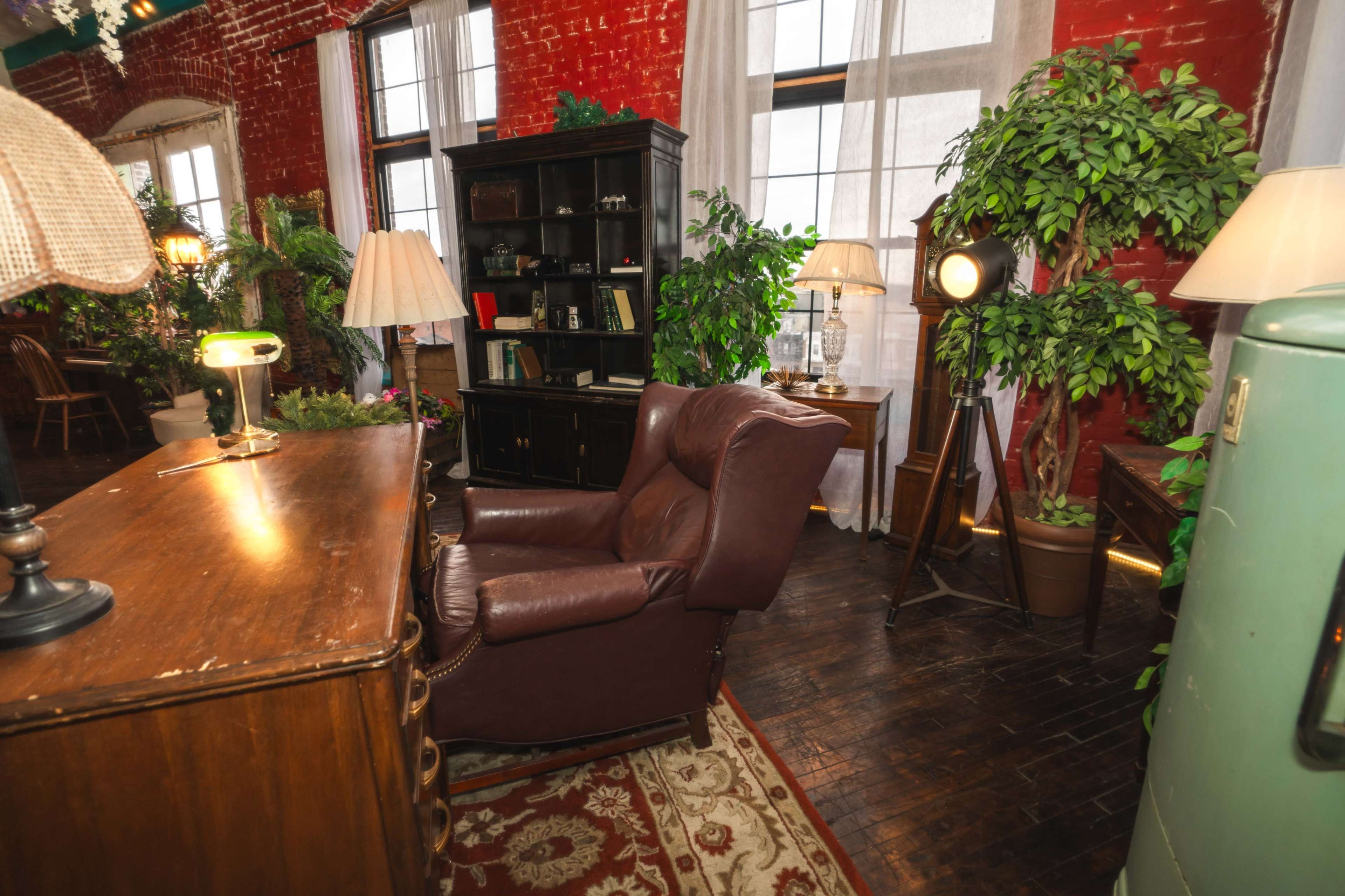 The room features a brown leather recliner next to a wooden desk, surrounded by plants and vintage decor against a backdrop of exposed red brick walls.