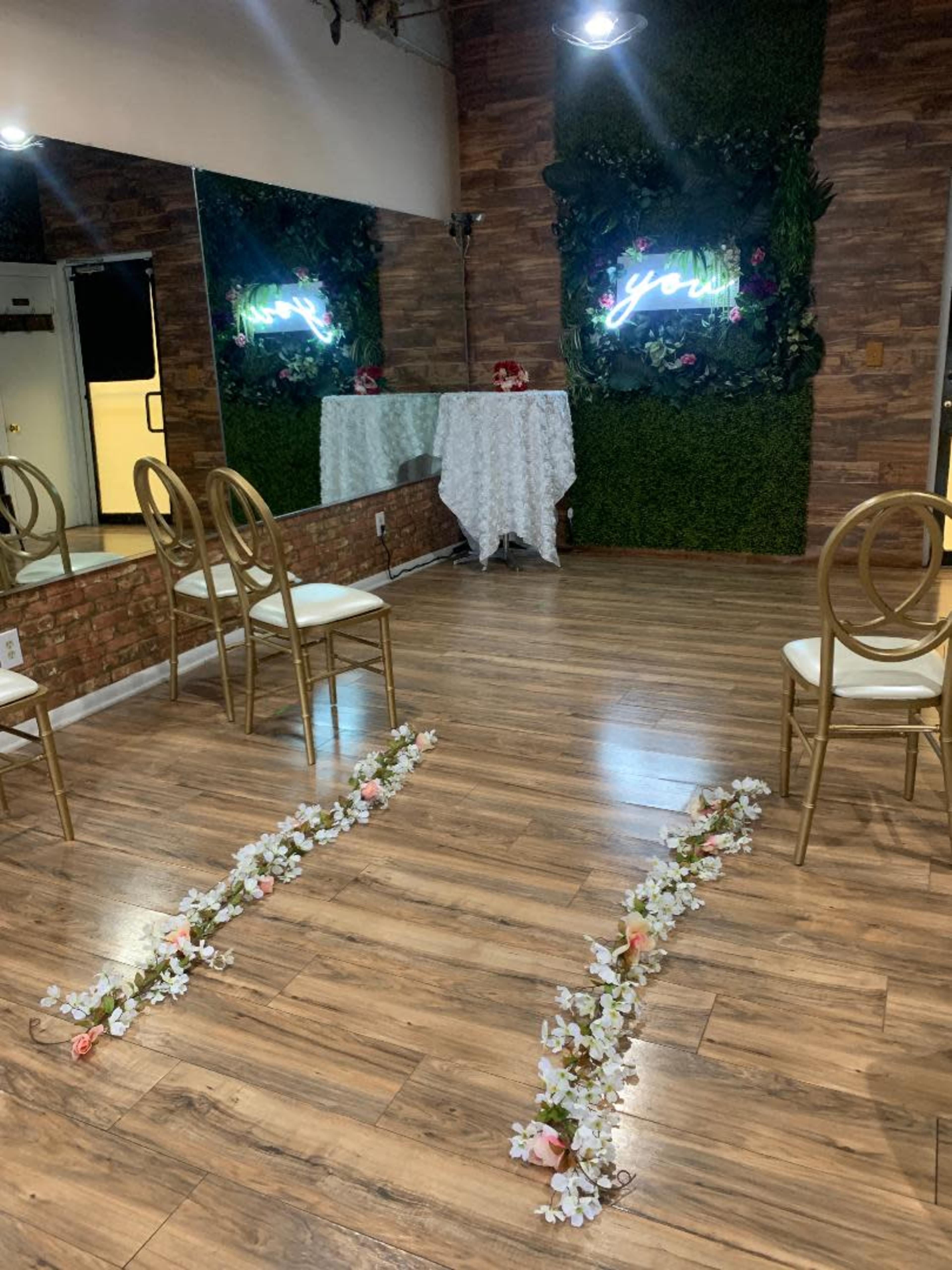 A decorated indoor space set up for an event, featuring gold chairs, a floral path on the wooden floor, and a table draped with white cloth against a backdrop of greenery and neon signage.