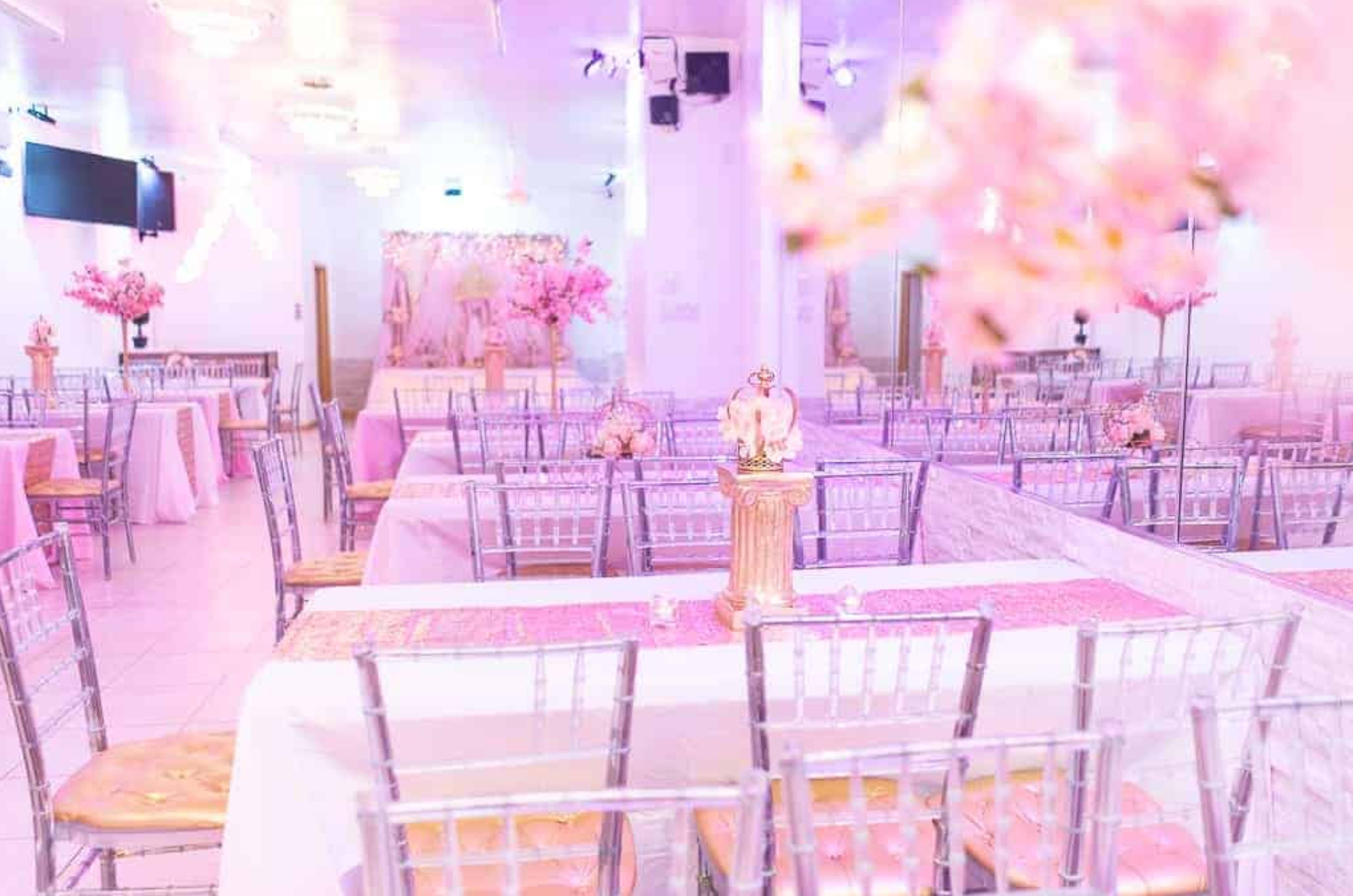 The image shows a decorated banquet hall with silver chiavari chairs, pink tablecloths, and floral centerpieces, set up for an event.