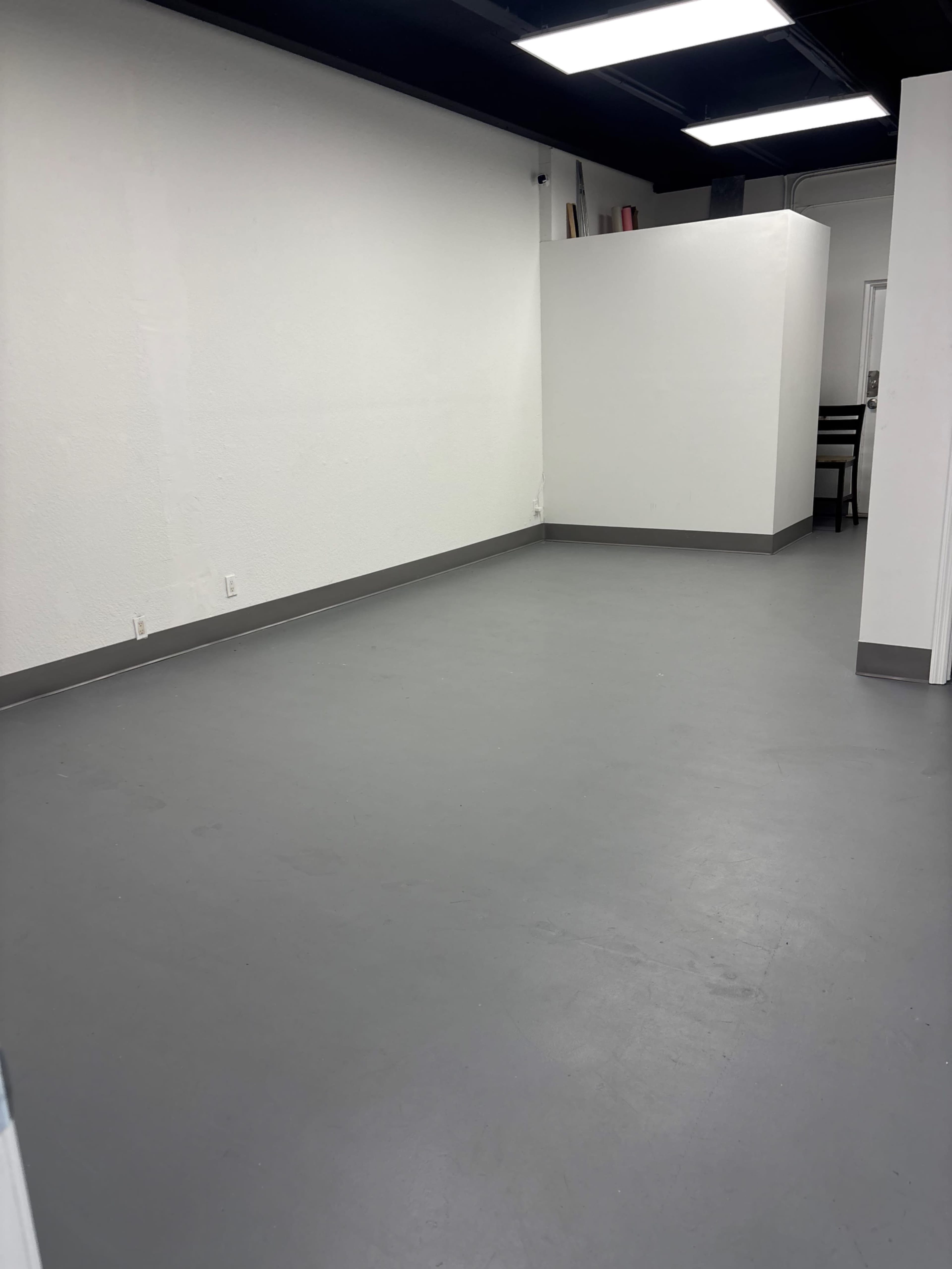 The image shows a sparsely furnished, empty room with grey flooring and white walls.