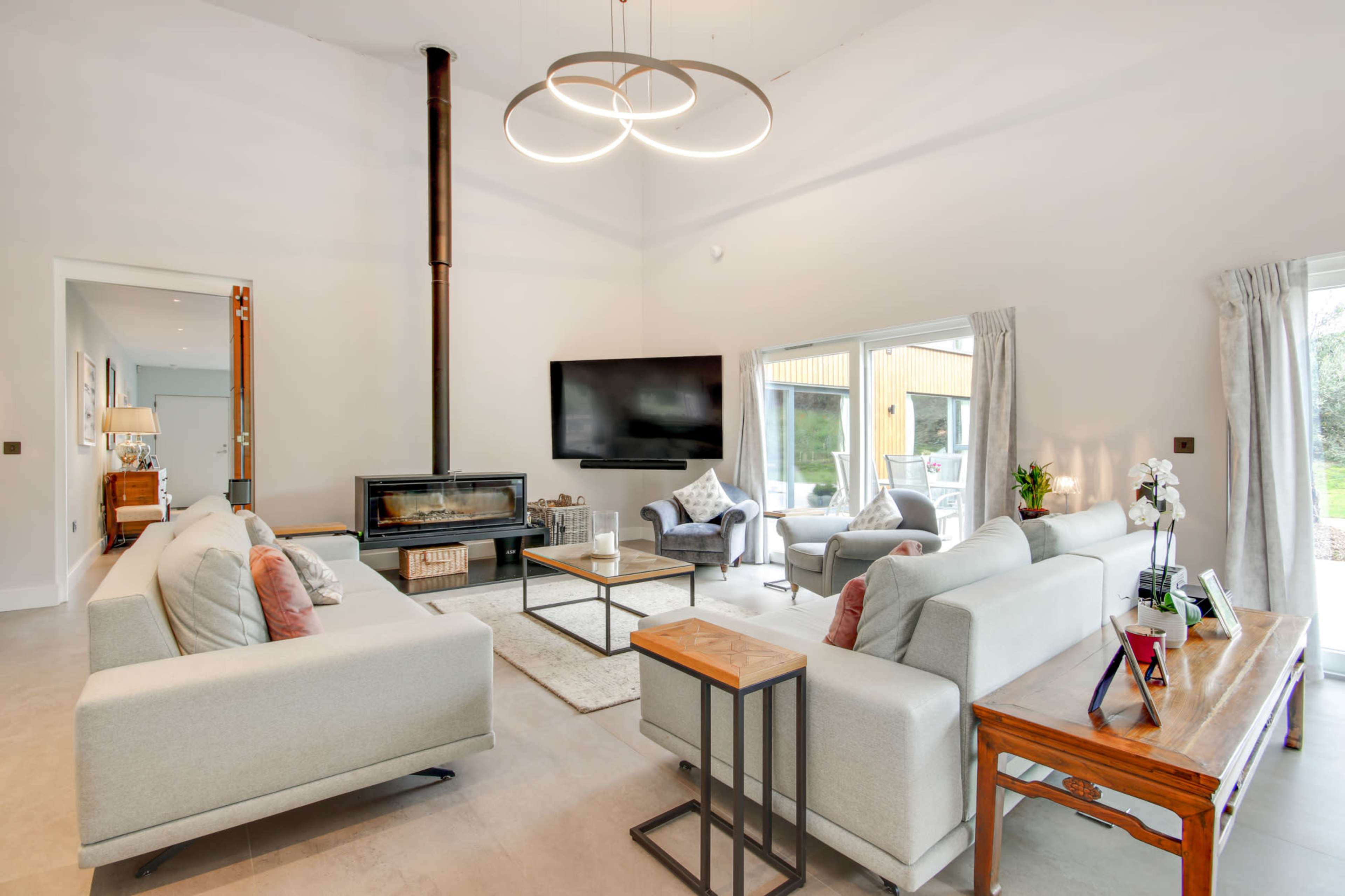 The image shows a modern living room featuring light-colored sofas, a television mounted on the wall, a fireplace, and large windows overlooking a garden.