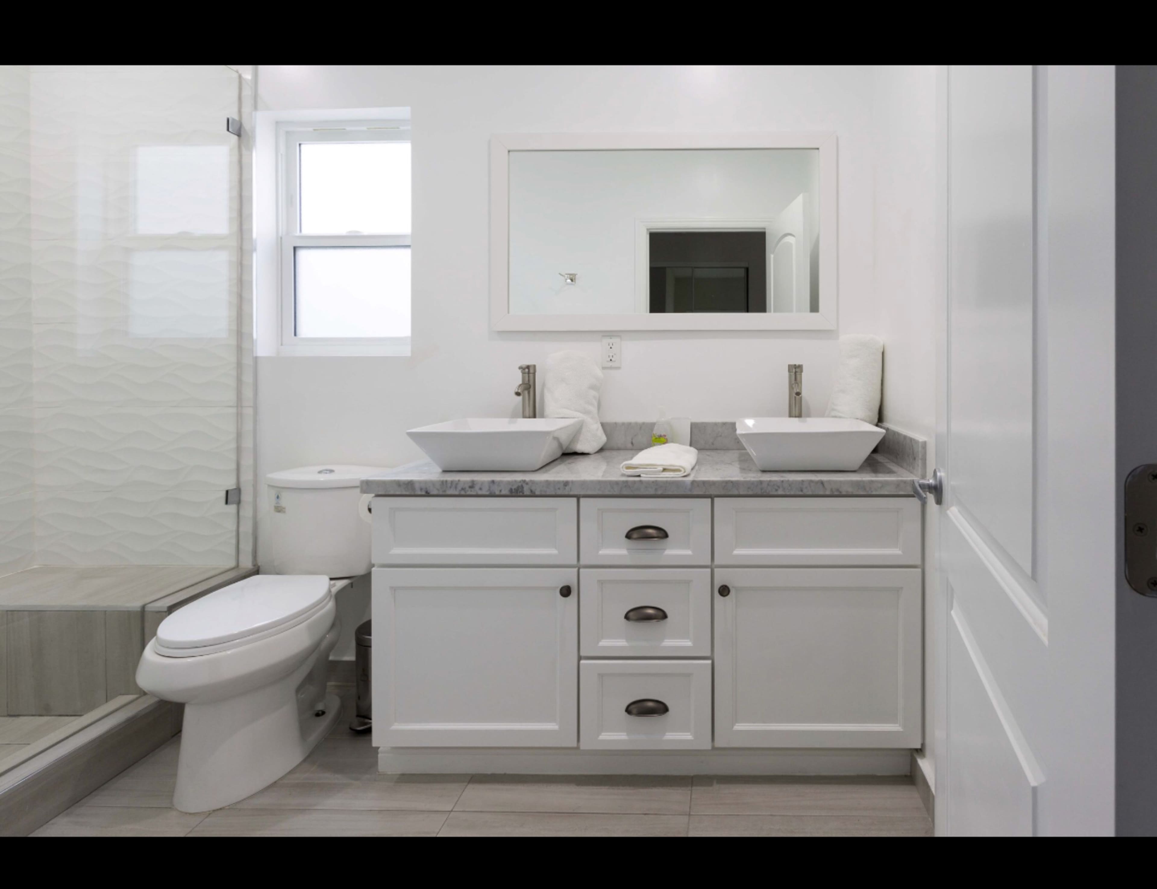 A modern bathroom with a white vanity, two sink basins, a large mirror, and a glass shower enclosure.
