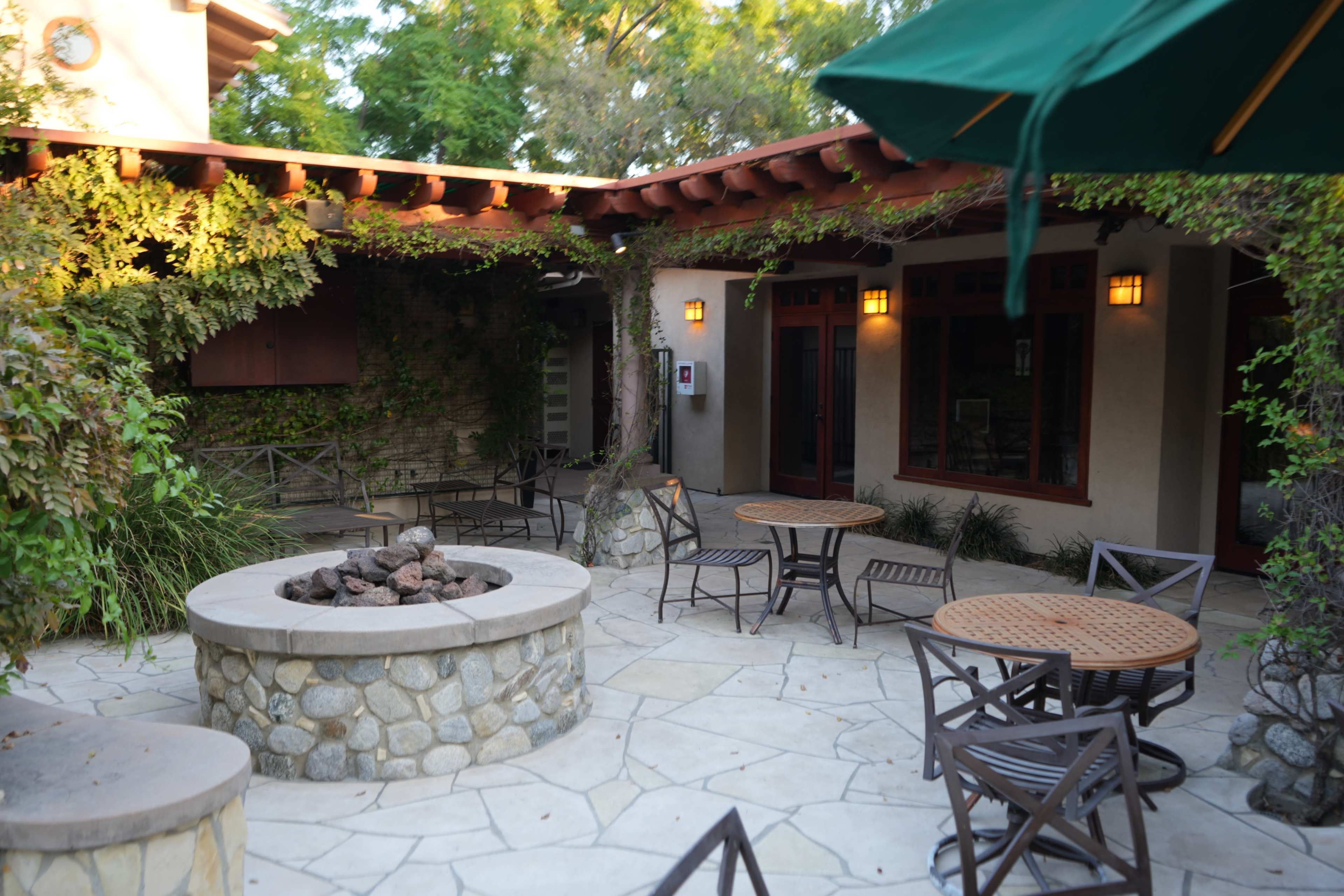 The image shows a landscaped outdoor courtyard with stone tables and chairs arranged around a circular fire pit, surrounded by greenery and walls of a building.