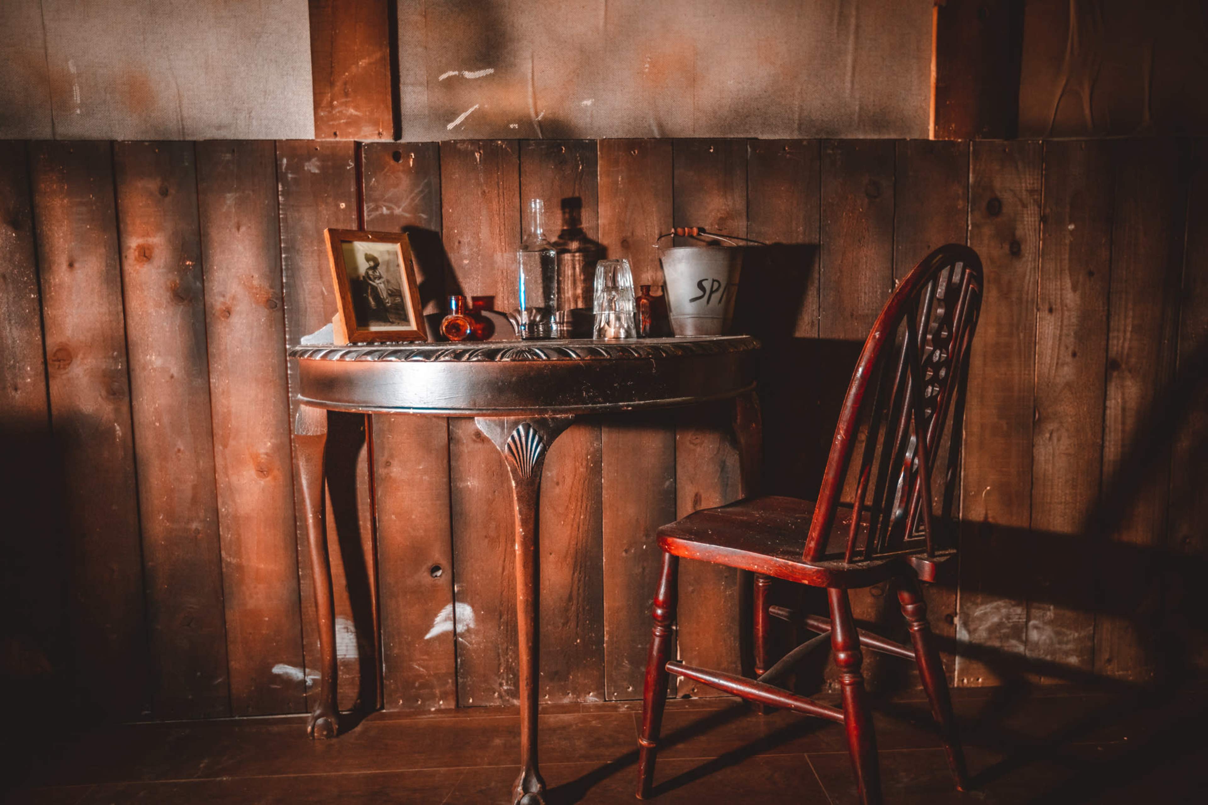 A wooden table with a decorative edge holds a bottle, glass, and small items, accompanied by a red chair against a wooden wall.
