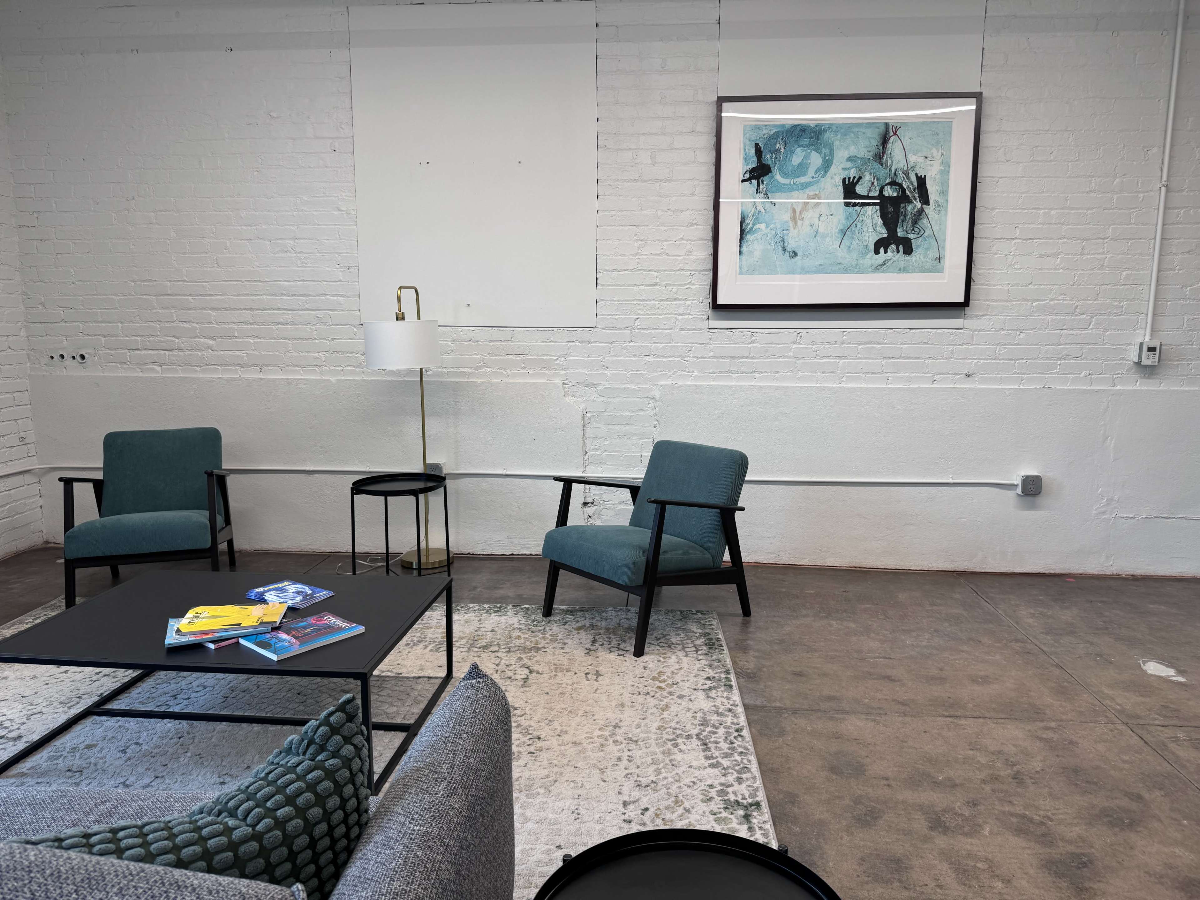 The image shows a minimalist room with two green chairs, a black side table, a gray sofa, and a framed artwork on the white brick wall.