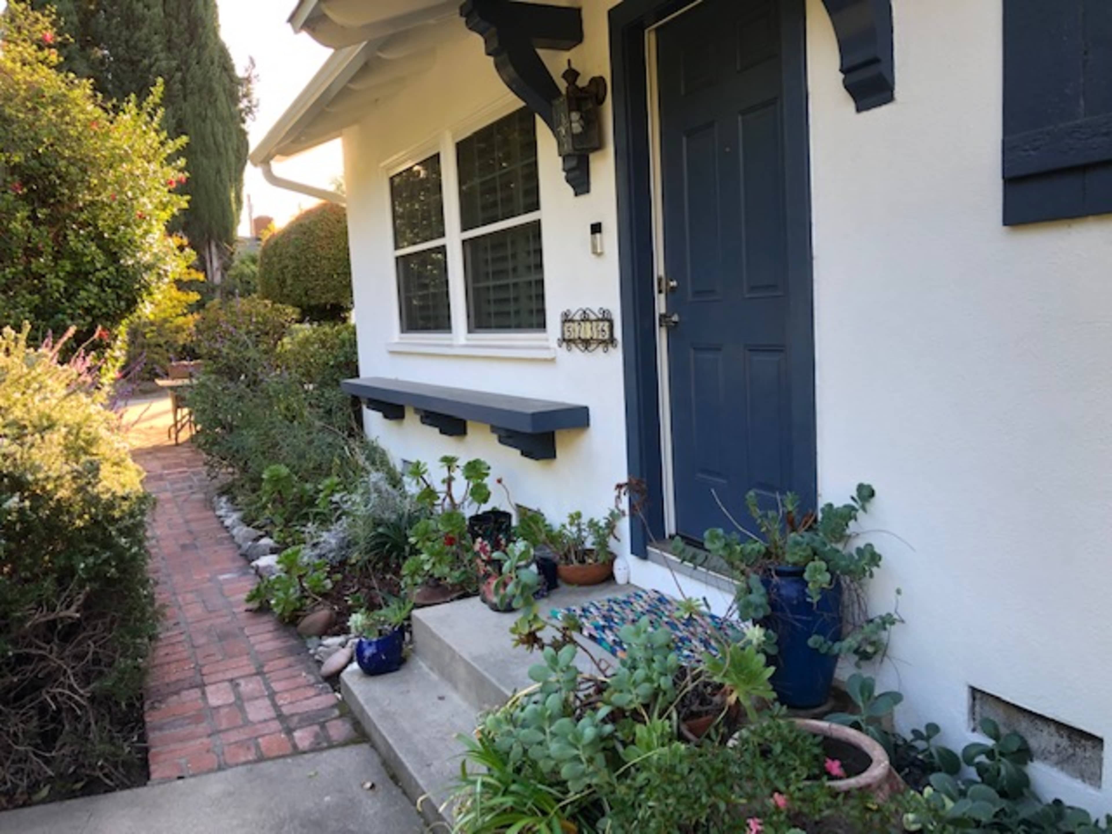 A pathway lined with potted plants leads to a blue front door of a house with white walls and decorative trim.