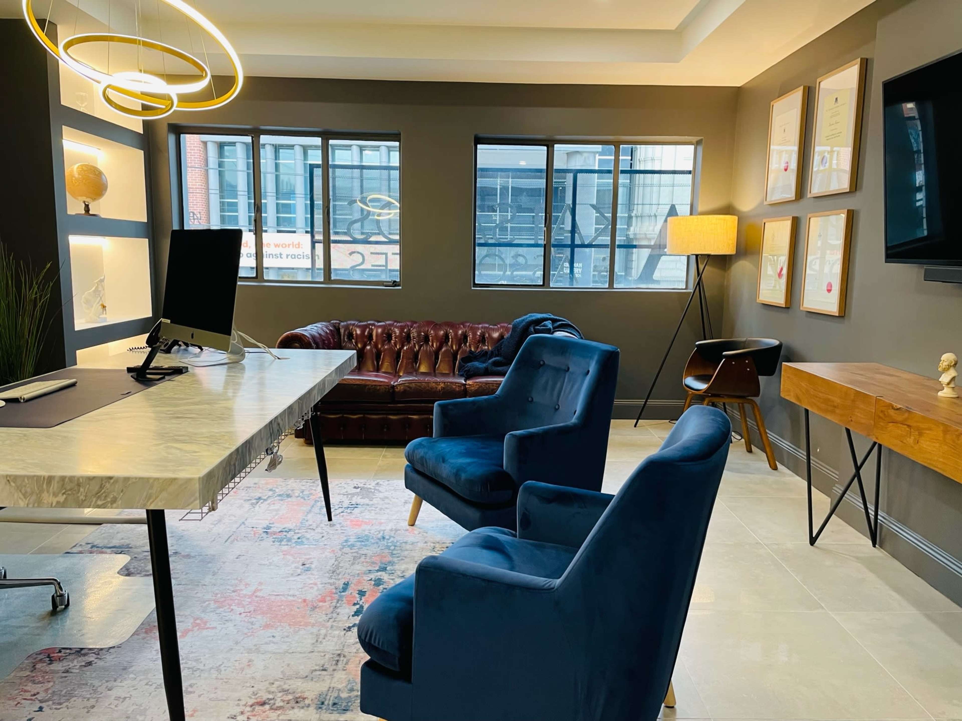 The image shows a modern office space featuring a marble-top desk, two blue chairs, a brown leather couch, a wooden side table, and framed certificates on the wall.