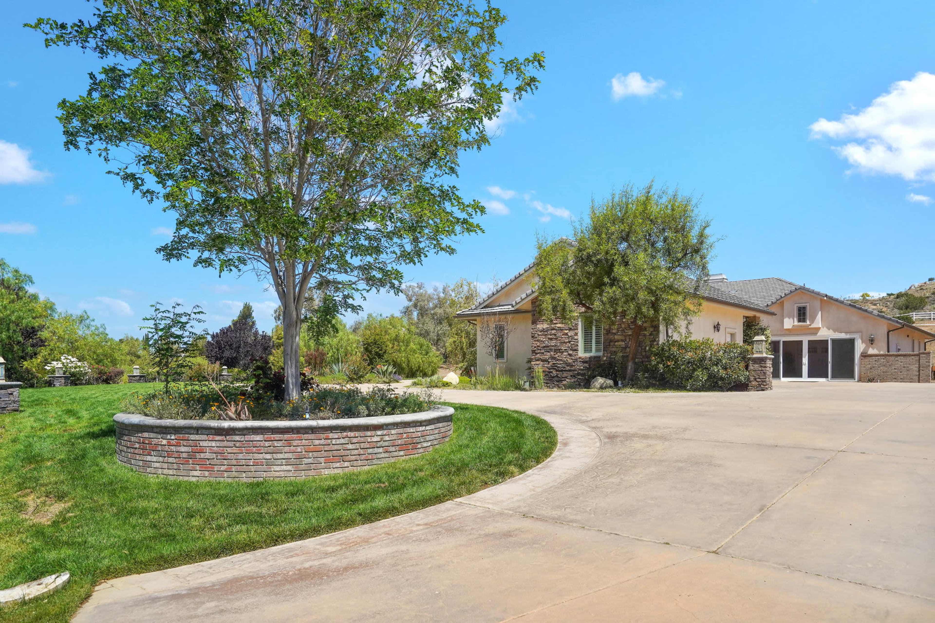 The image shows a residential property with a circular driveway, a landscaped yard featuring a tree and flower beds, and a house designed with stone accents.