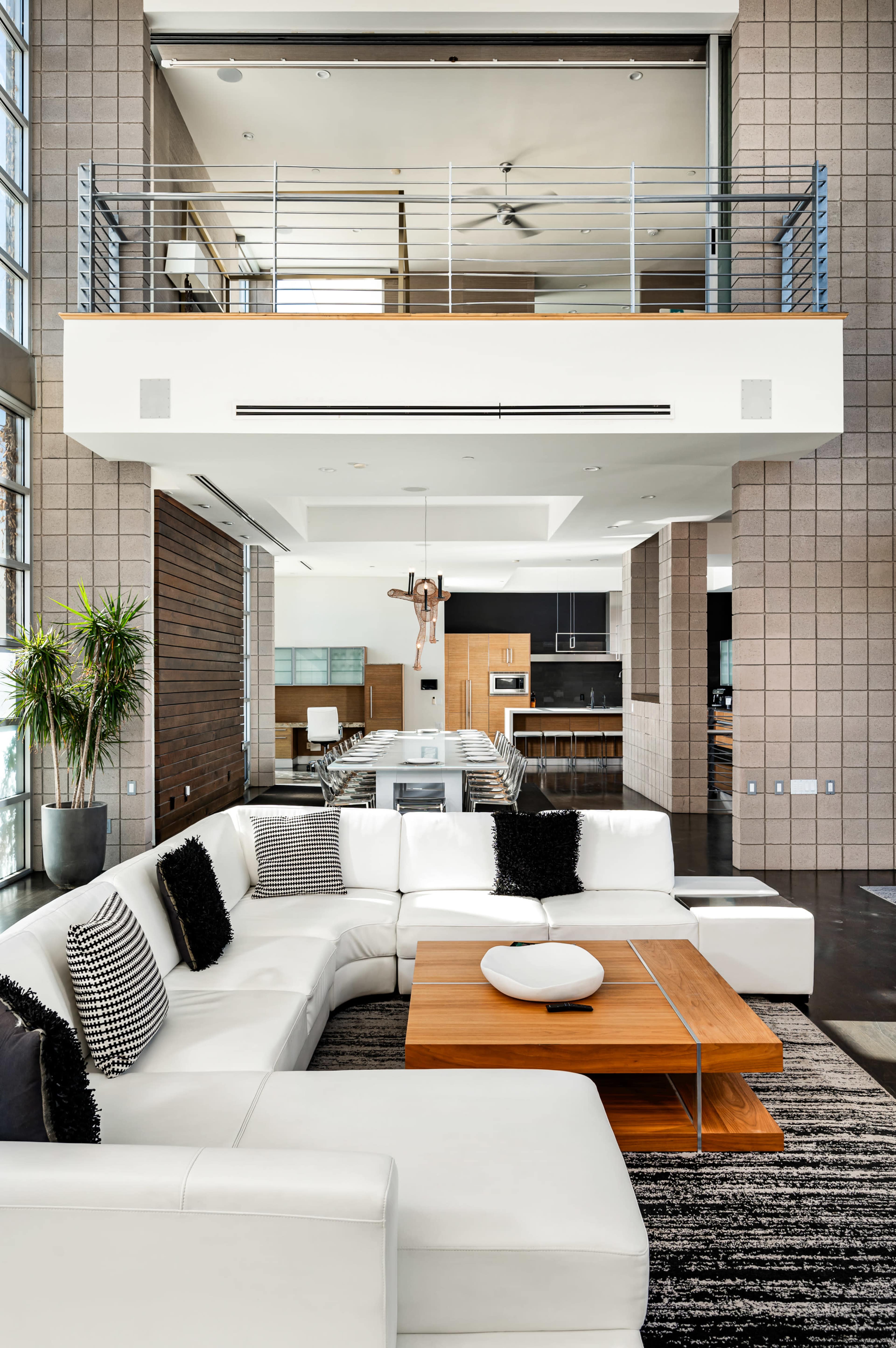 A modern, spacious living room with a large white sectional sofa and a wooden coffee table, overlooking an open kitchen area on a higher level.