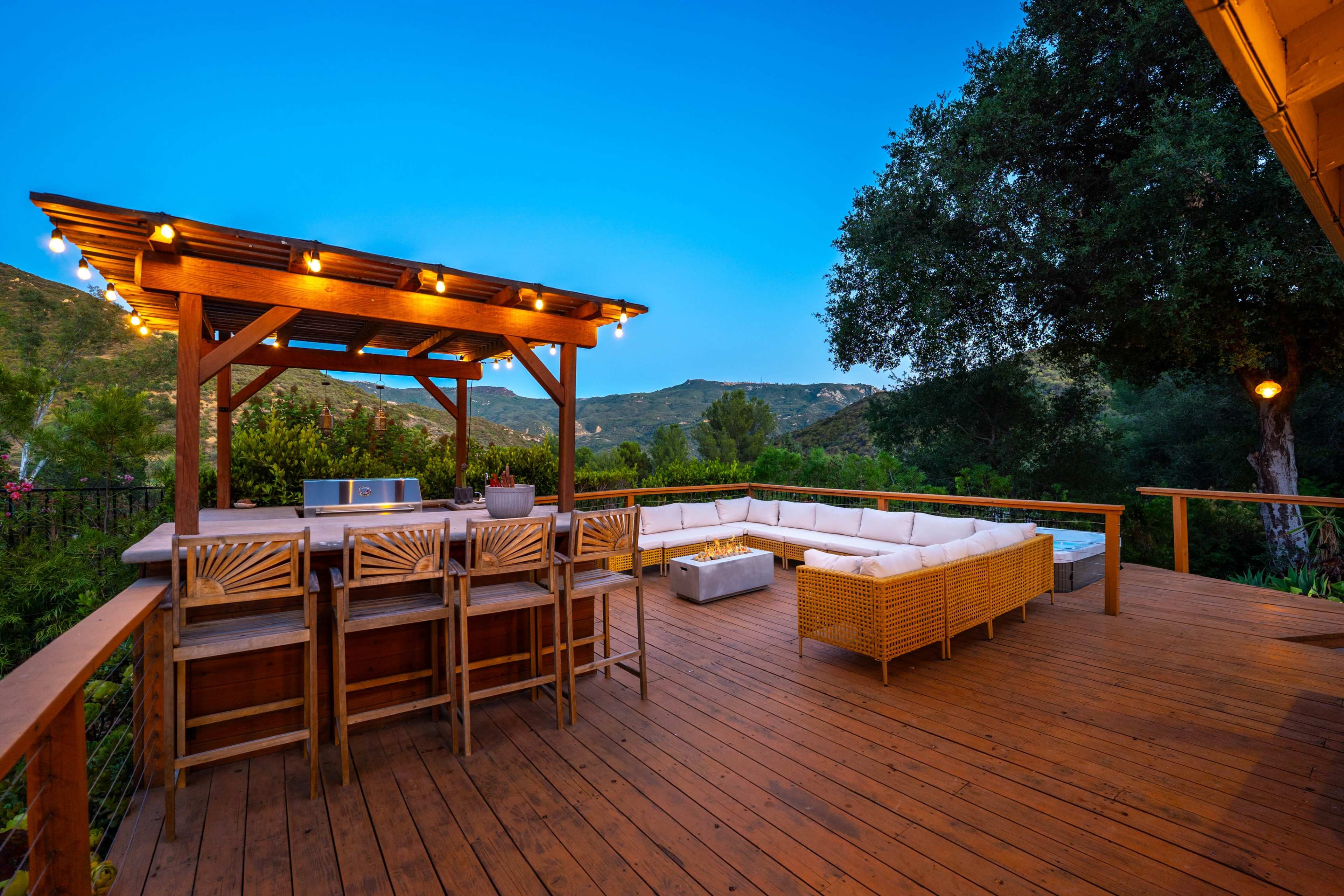 The image shows a wooden deck featuring a bar with seating, a cushioned lounge area, and a backdrop of rolling hills under a twilight sky.
