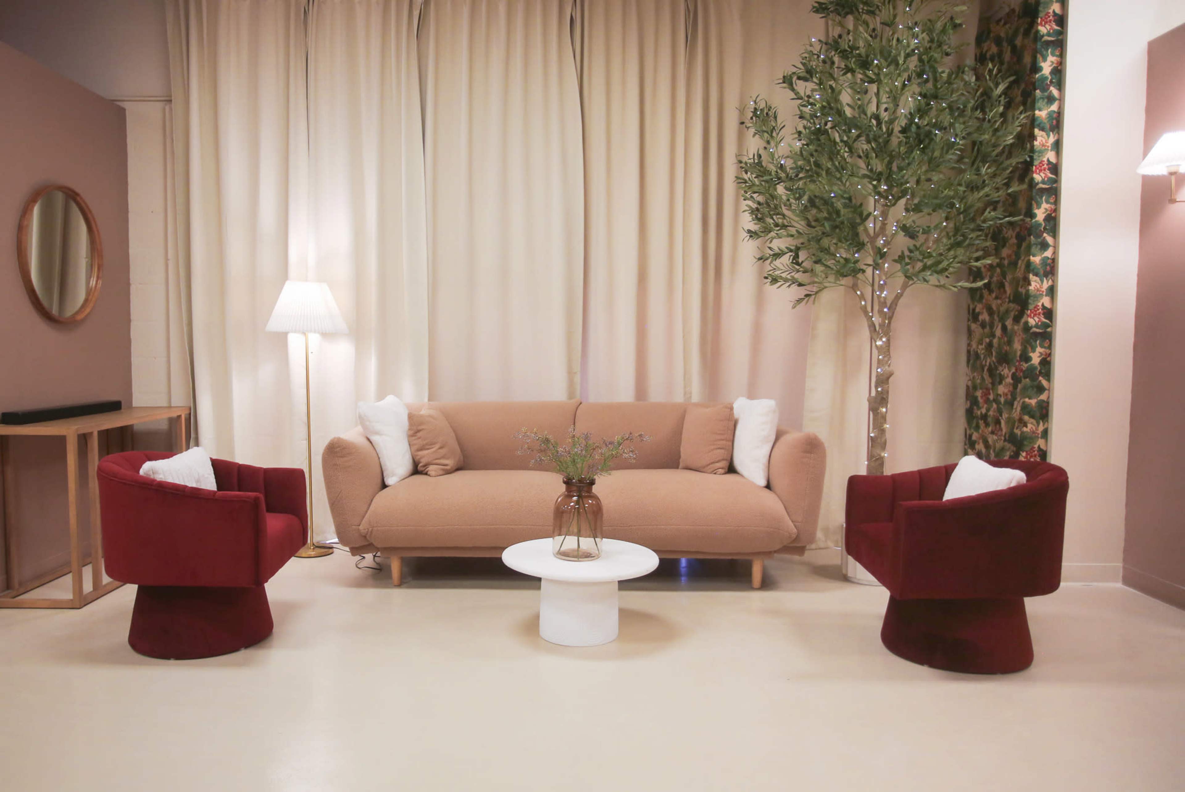A minimalist living room with a beige sofa, two red armchairs, a round white coffee table, a floor lamp, and a decorative tree.