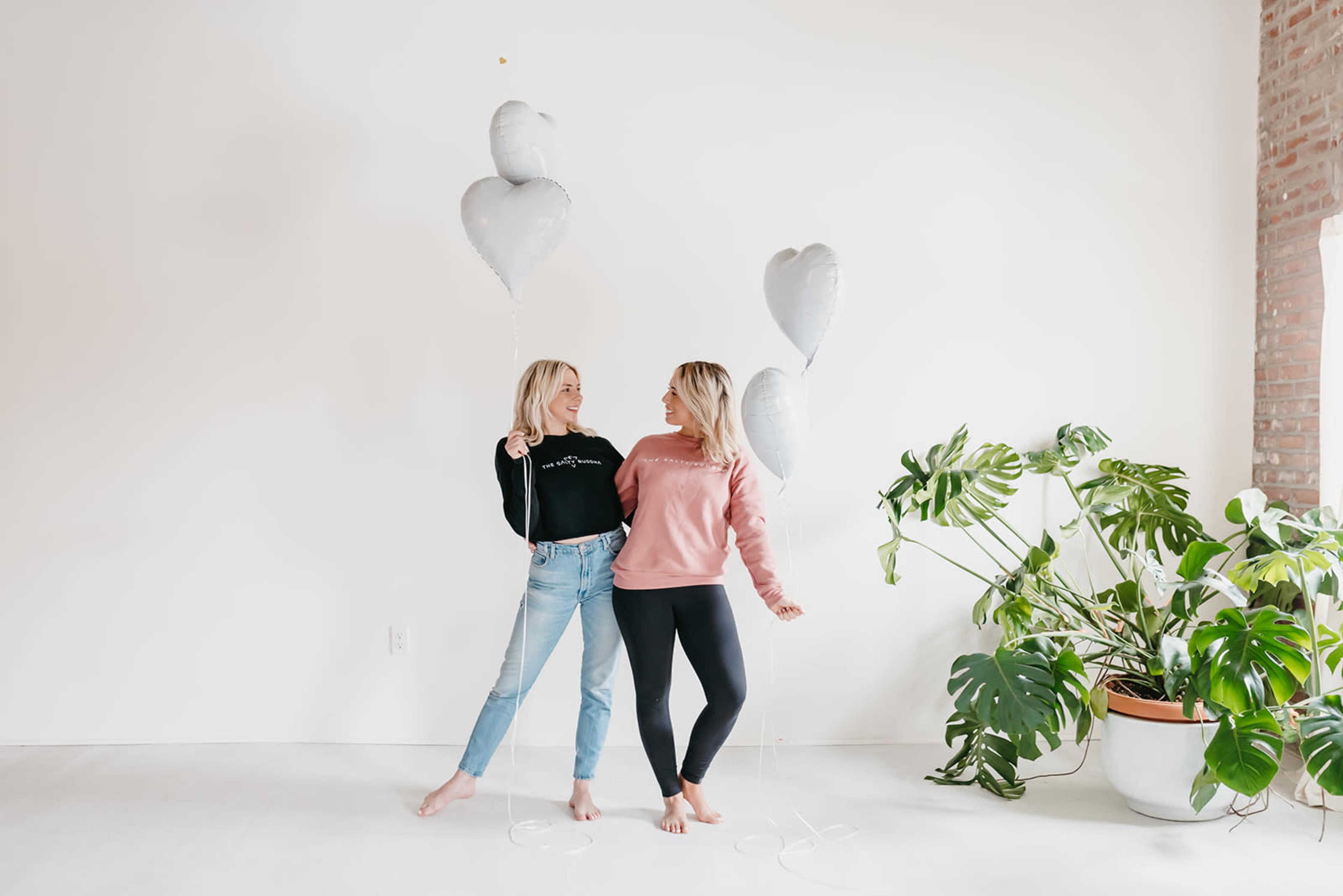 Two women stand in a bright room holding heart-shaped balloons, surrounded by a large potted plant.
