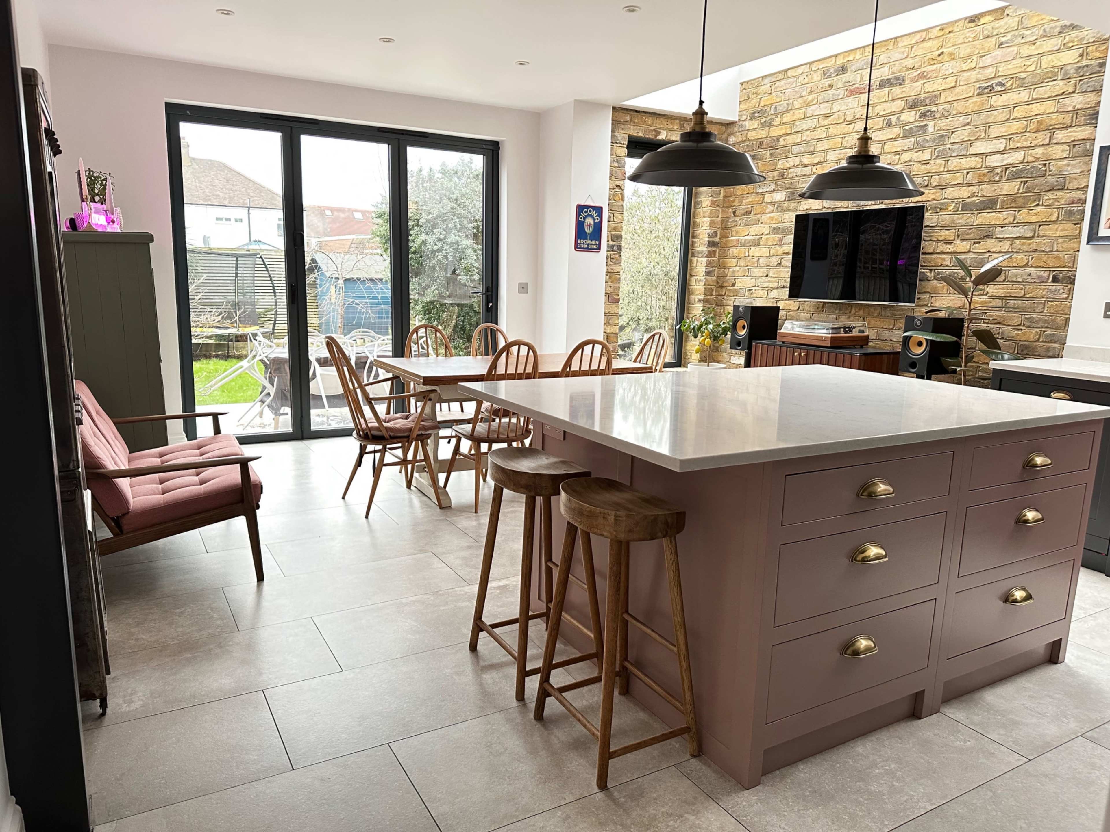 The image shows a modern kitchen with a large central island, wooden dining table, and a view of a backyard through large glass doors.