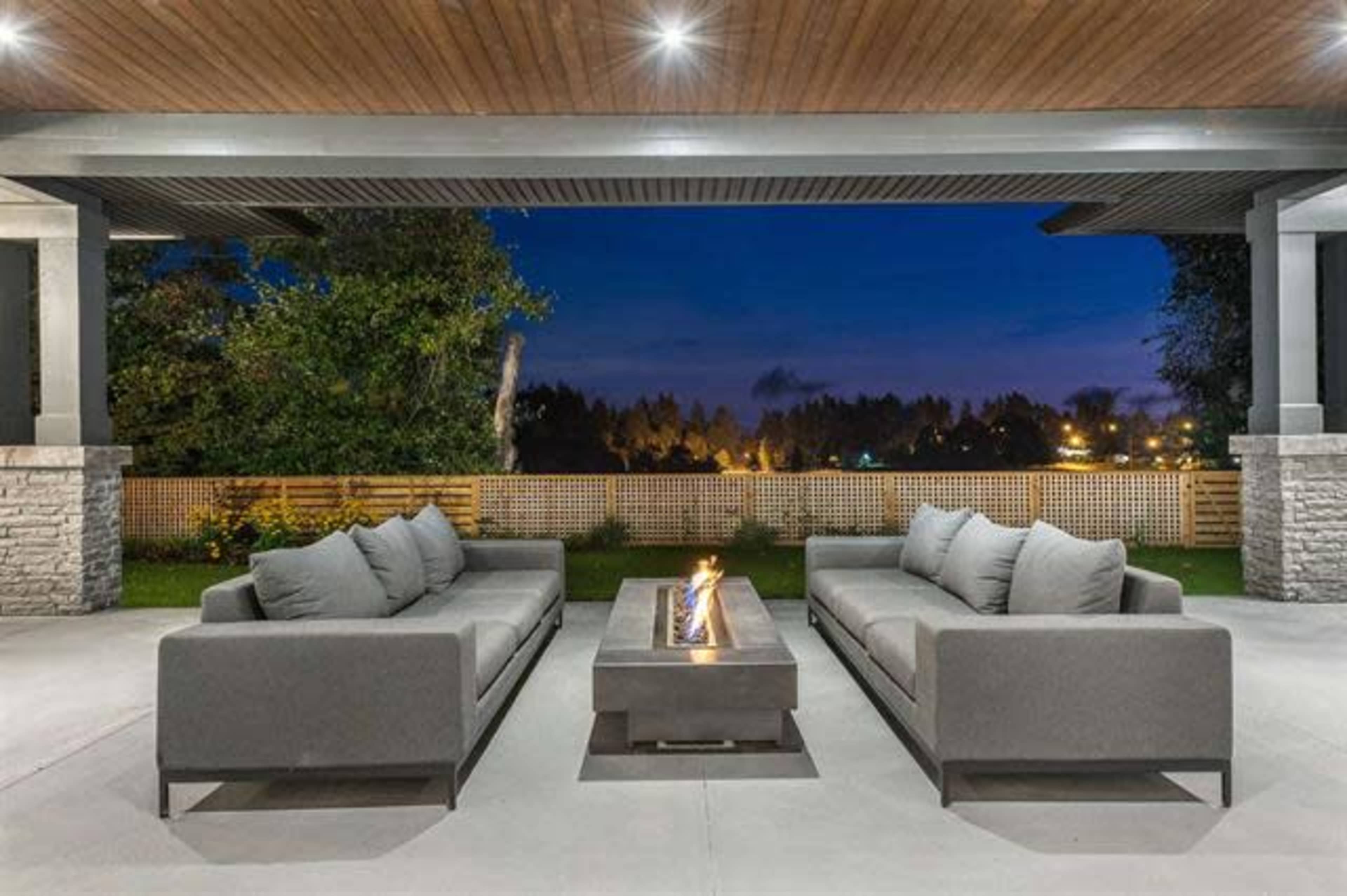 A modern outdoor seating area features two gray sectional sofas arranged around a central fire pit, set against a backdrop of trees and dim evening sky.