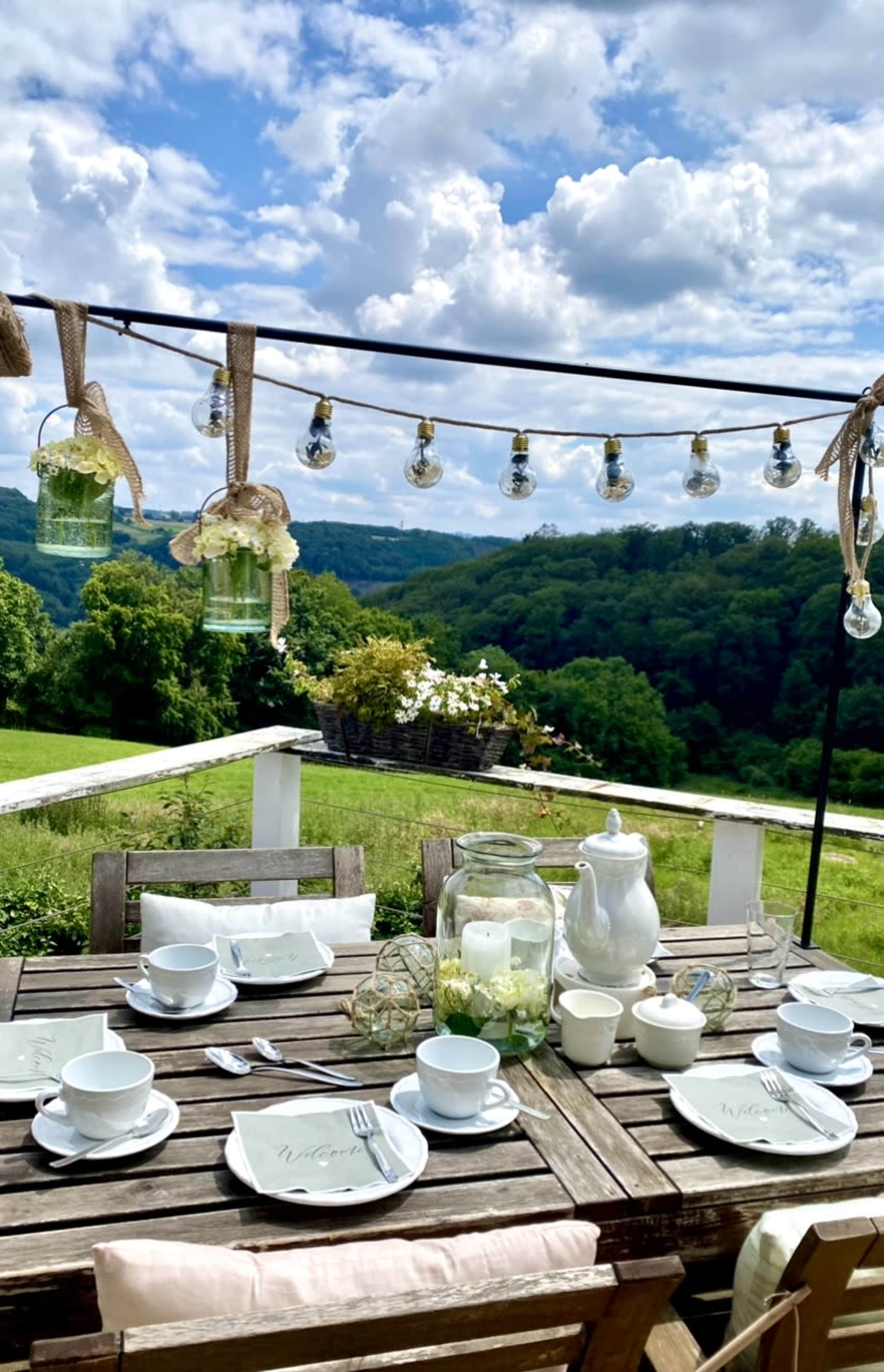 A wooden table is set for a meal outdoors, adorned with dishes, glasses, and decorative lanterns, overlooking a lush green landscape under a cloudy sky.