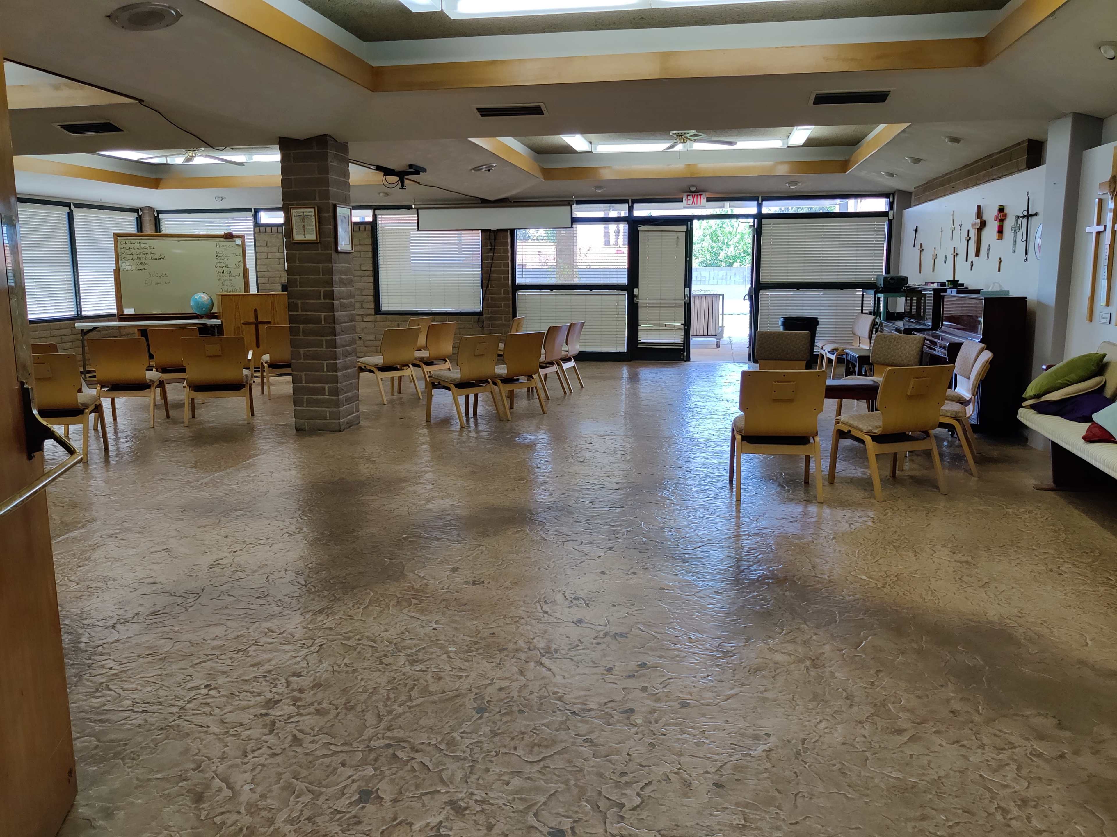 A spacious indoor room with chairs arranged around the area, a piano against the wall, and various crosses displayed, along with a large door leading outside.