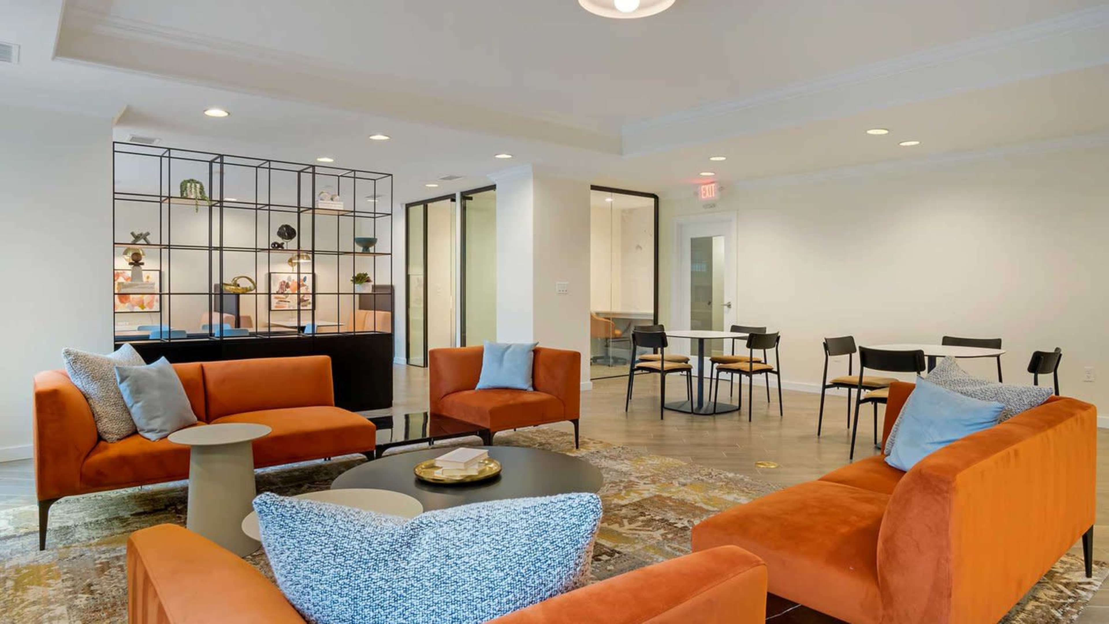 A modern lounge area featuring orange sofas, a coffee table, and a decorative shelving unit, with a dining area visible in the background.