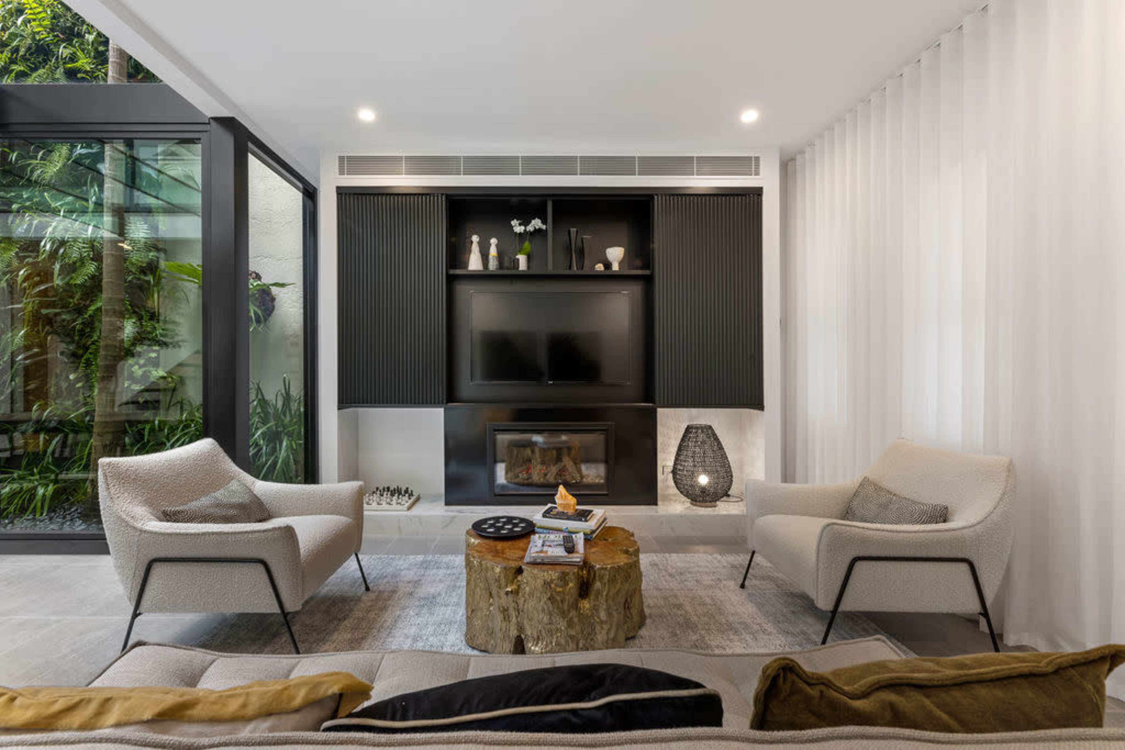 A modern living room with two armchairs, a wooden coffee table, a mounted television, and large windows allowing natural light.
