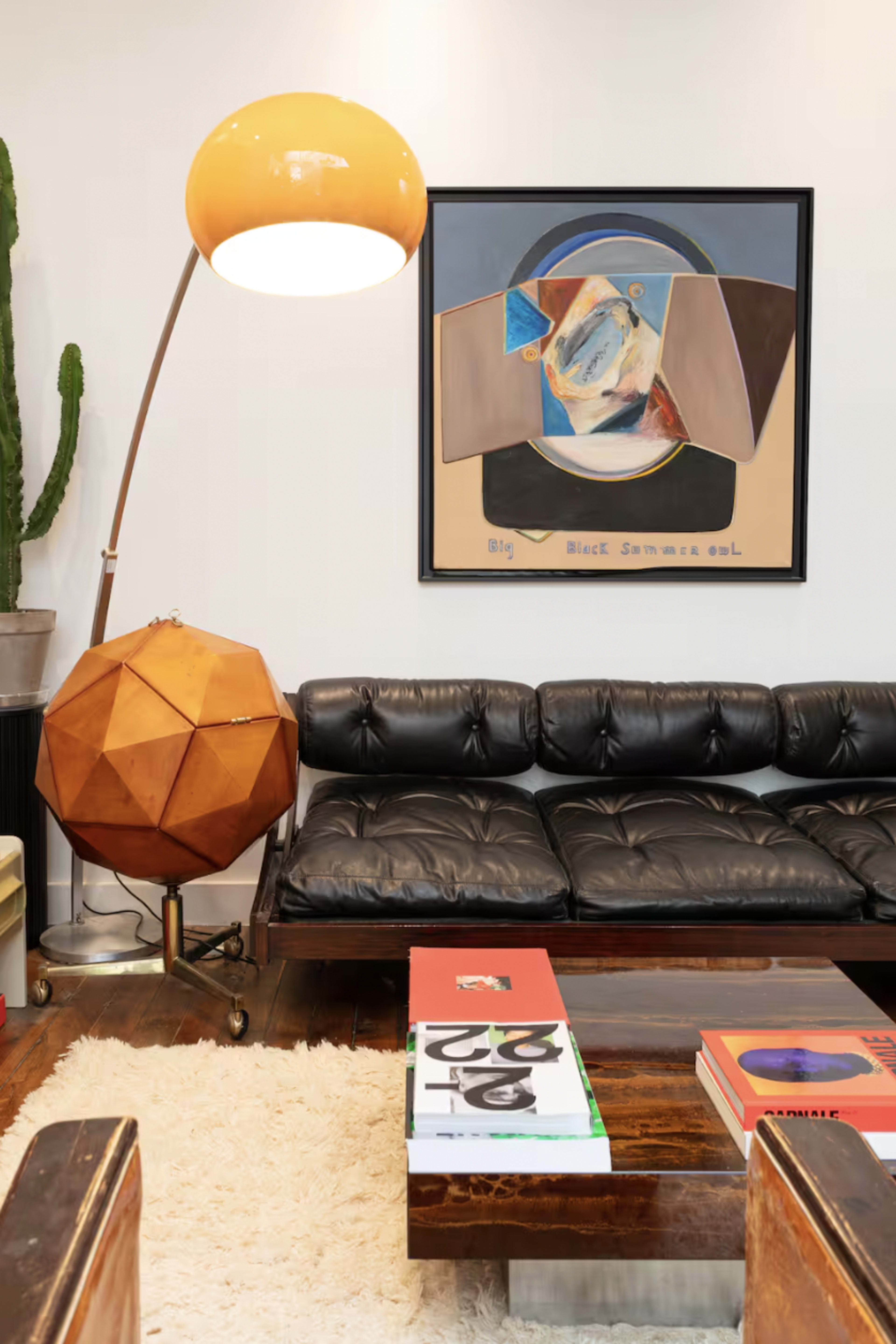 The image features a modern living room with a black leather sofa, a geometric floor lamp, a wooden coffee table stacked with books, and a large abstract painting on the wall.