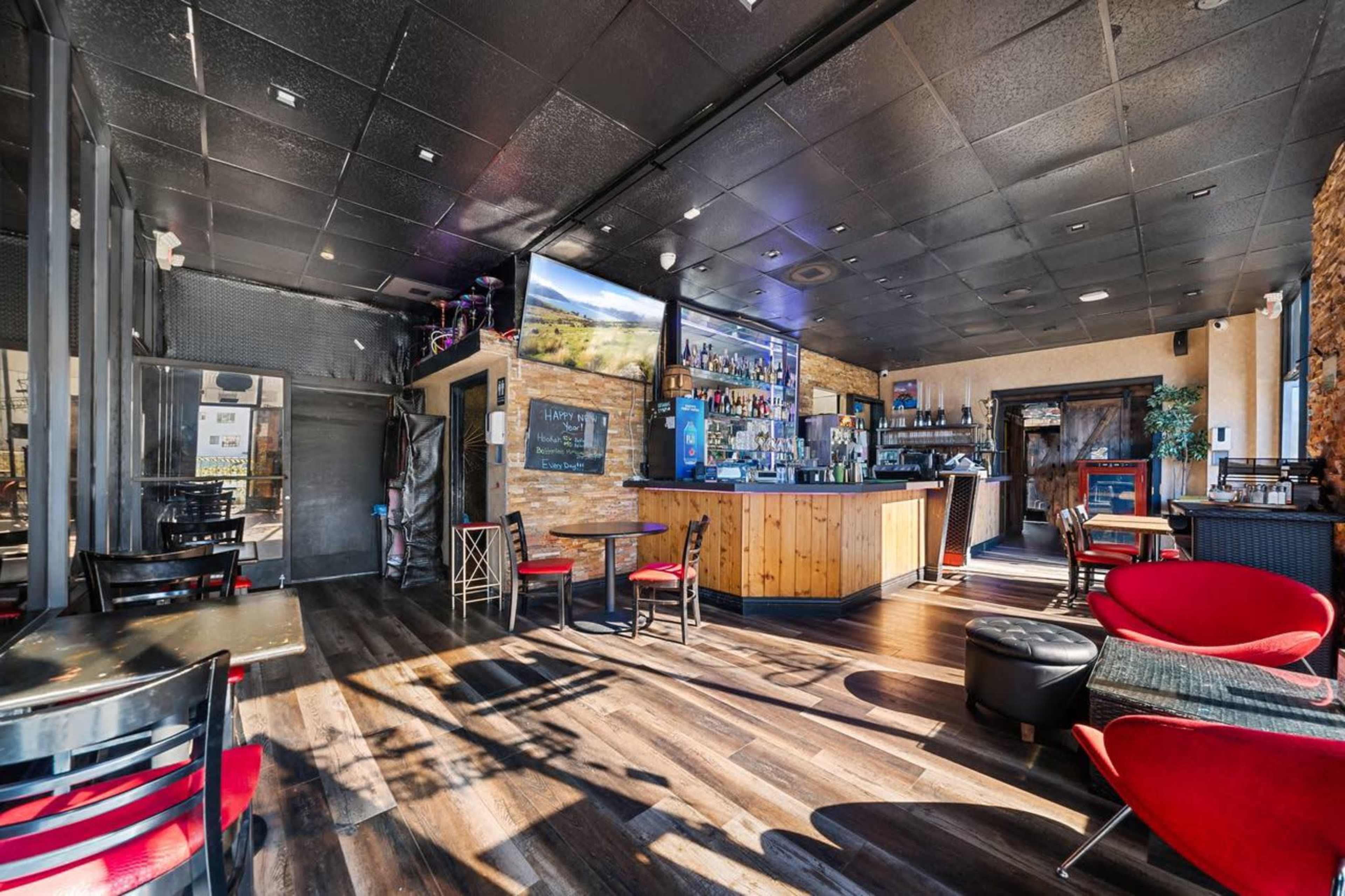 The image shows a modern bar interior with wooden flooring, a well-stocked bar counter, and chairs arranged around small tables.