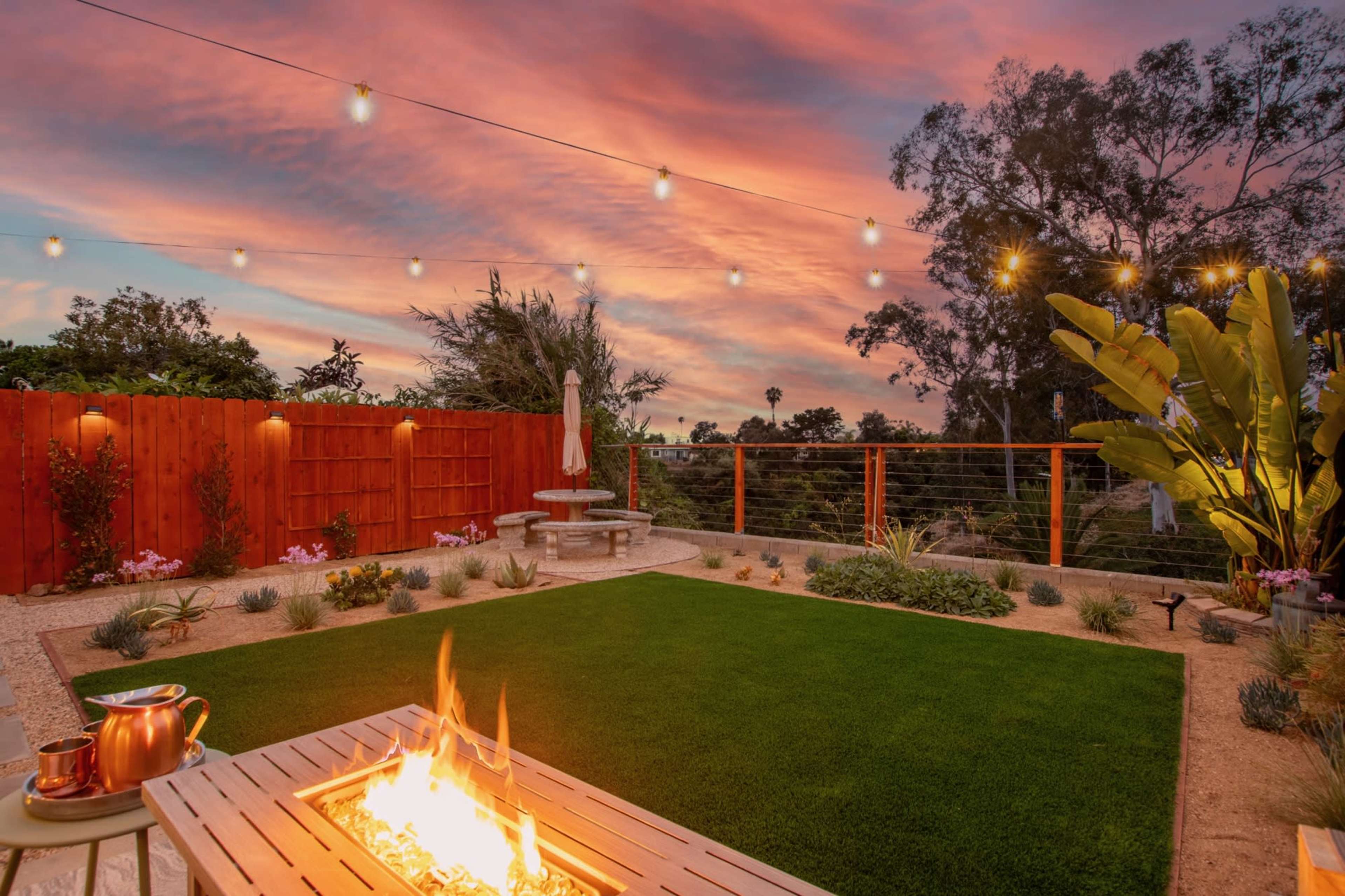 A landscaped backyard with a fire pit, green grass, and decorative string lights above at sunset.
