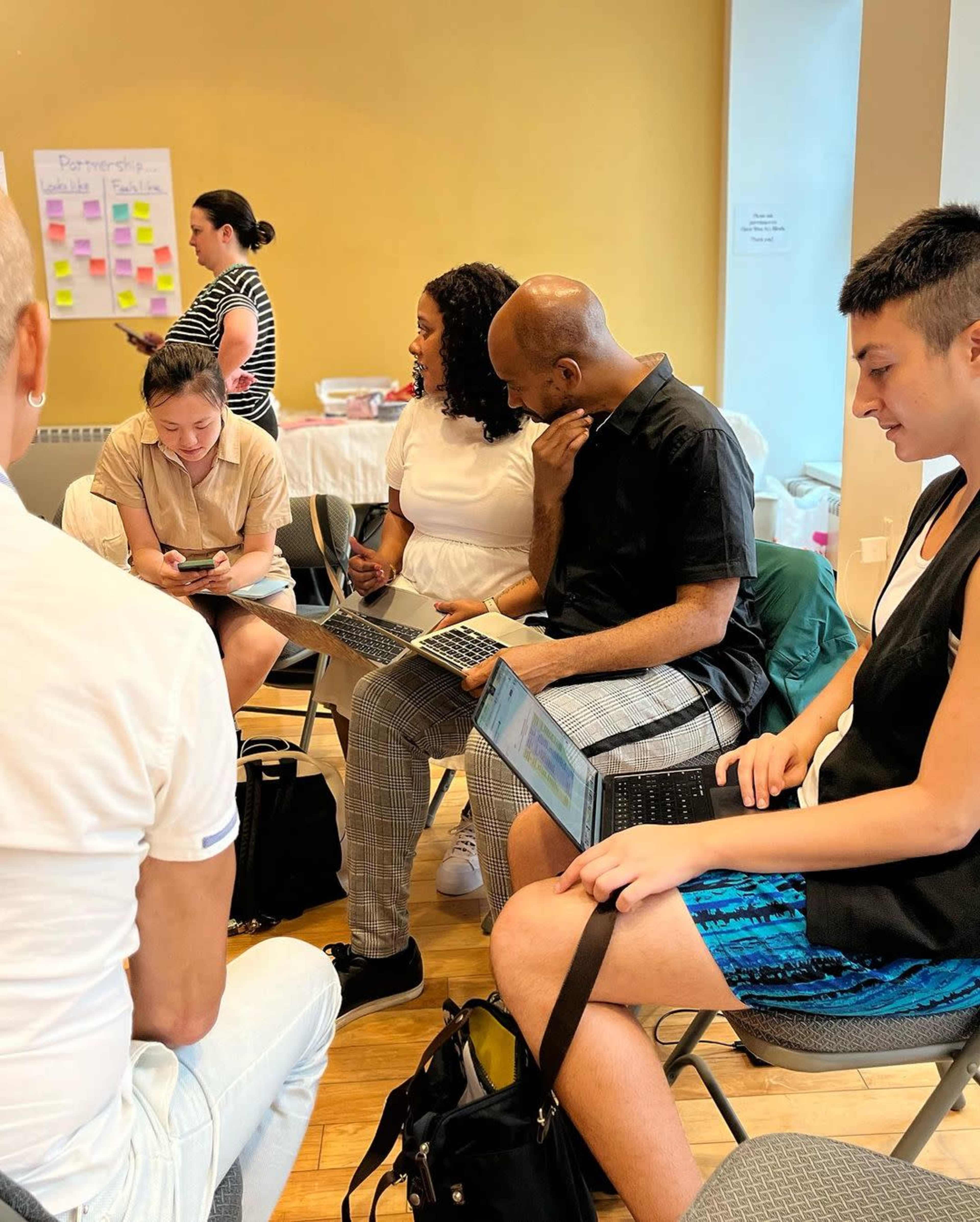A group of people sits in a circle, engaged with laptops and mobile devices in a collaborative workspace.
