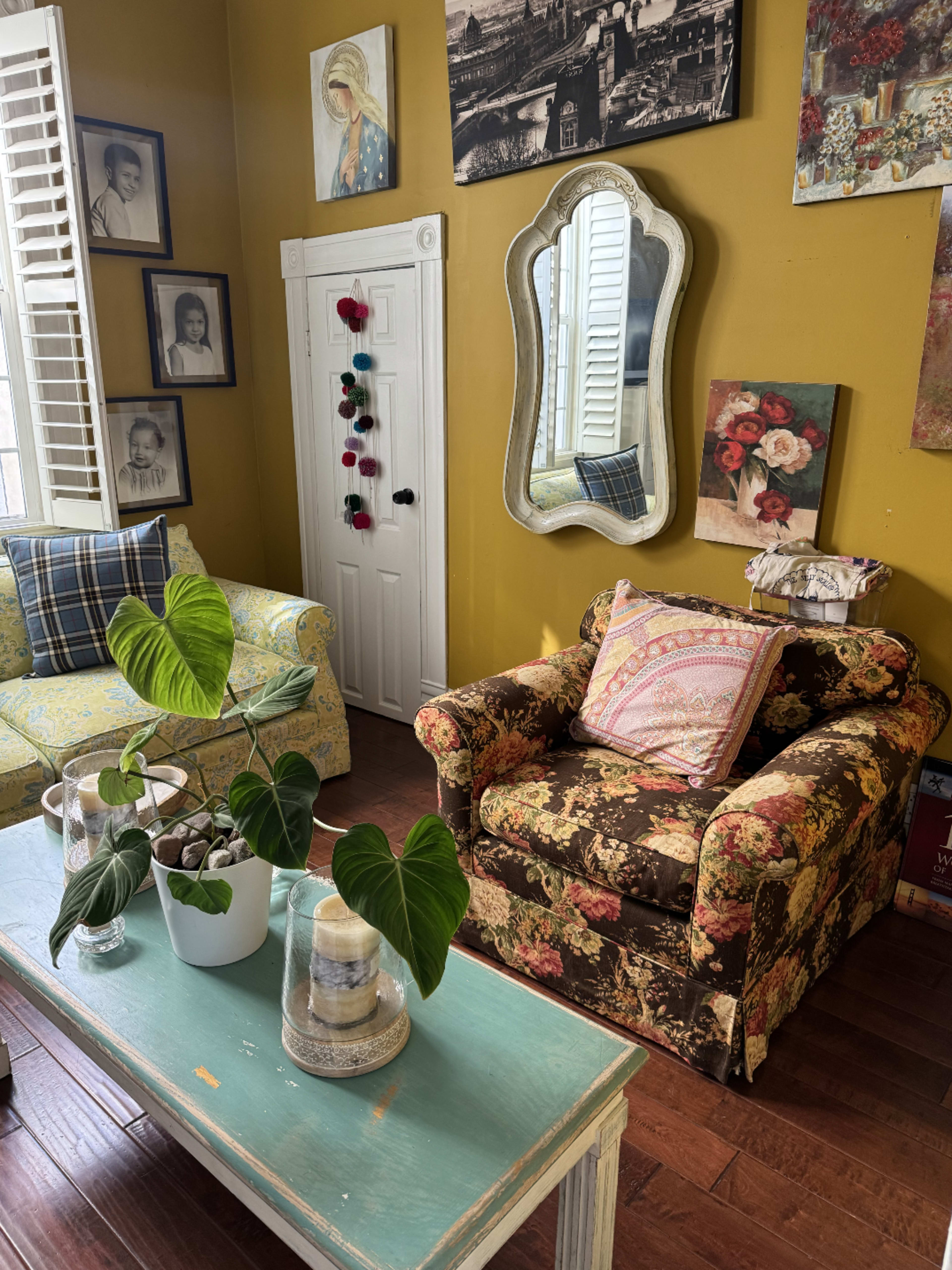 The room features two upholstered chairs, a wooden coffee table with decorative items, and various framed artworks on the yellow walls.