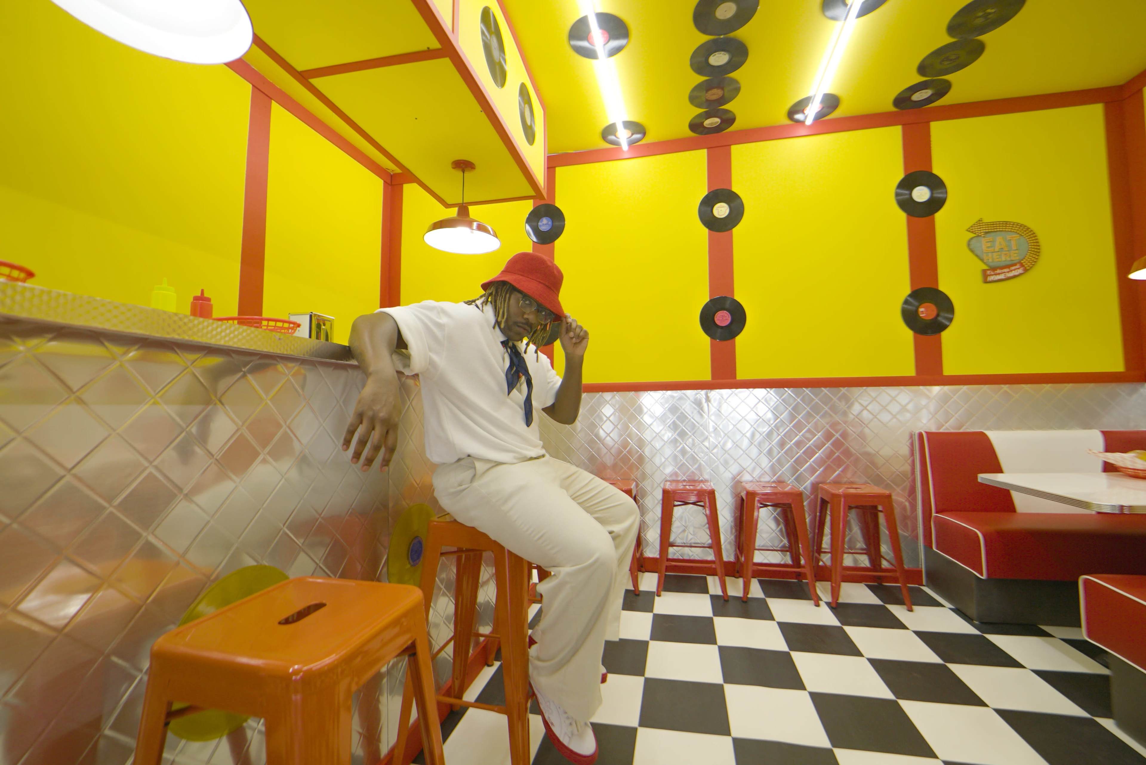 A person wearing a red hat and white outfit sits on a red stool in a brightly colored diner decorated with yellow walls and vintage record discs.
