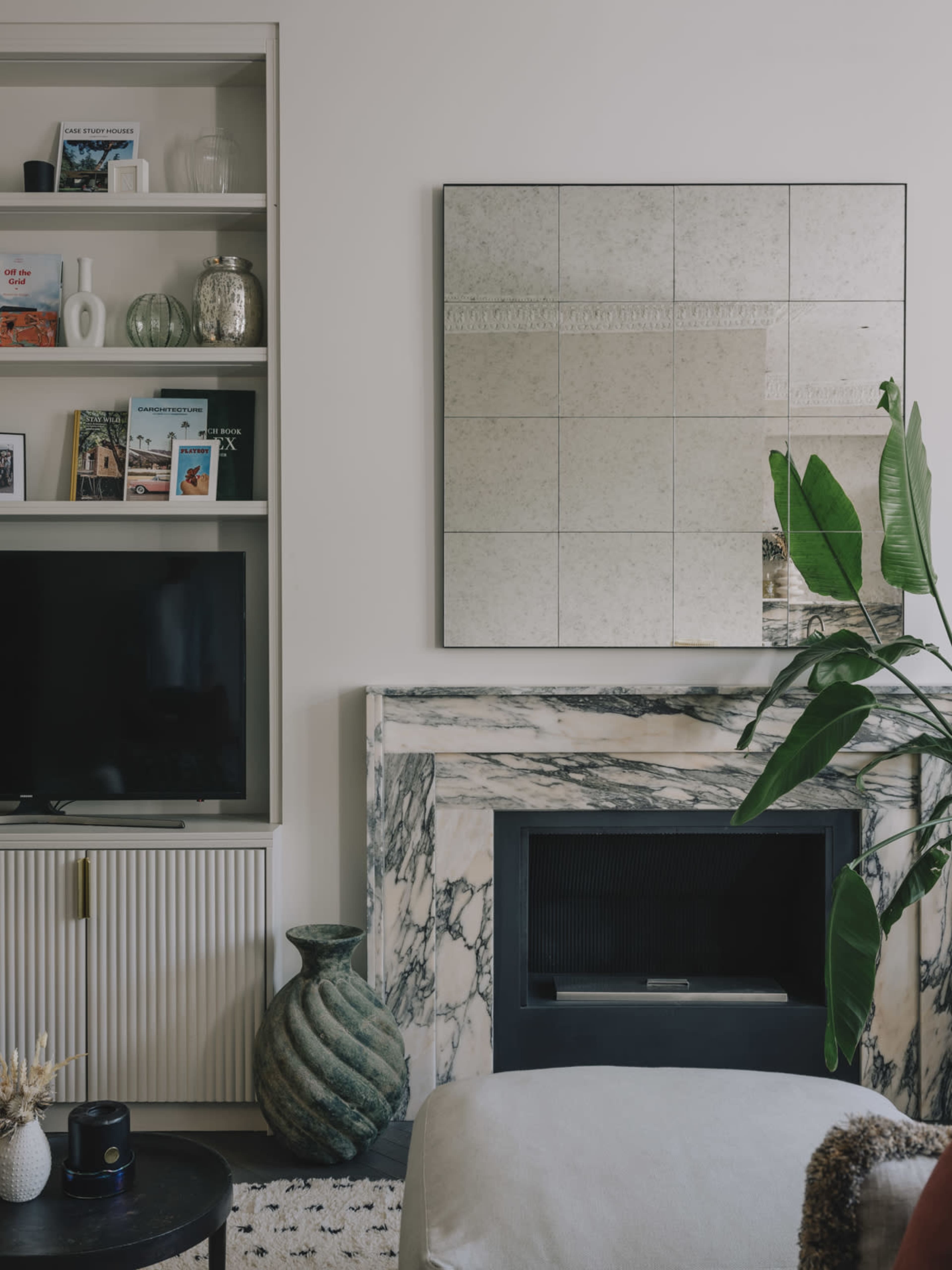 The image shows a modern living room featuring a marble fireplace, a large mirror, a television, and a bookshelf filled with decorative items and books.