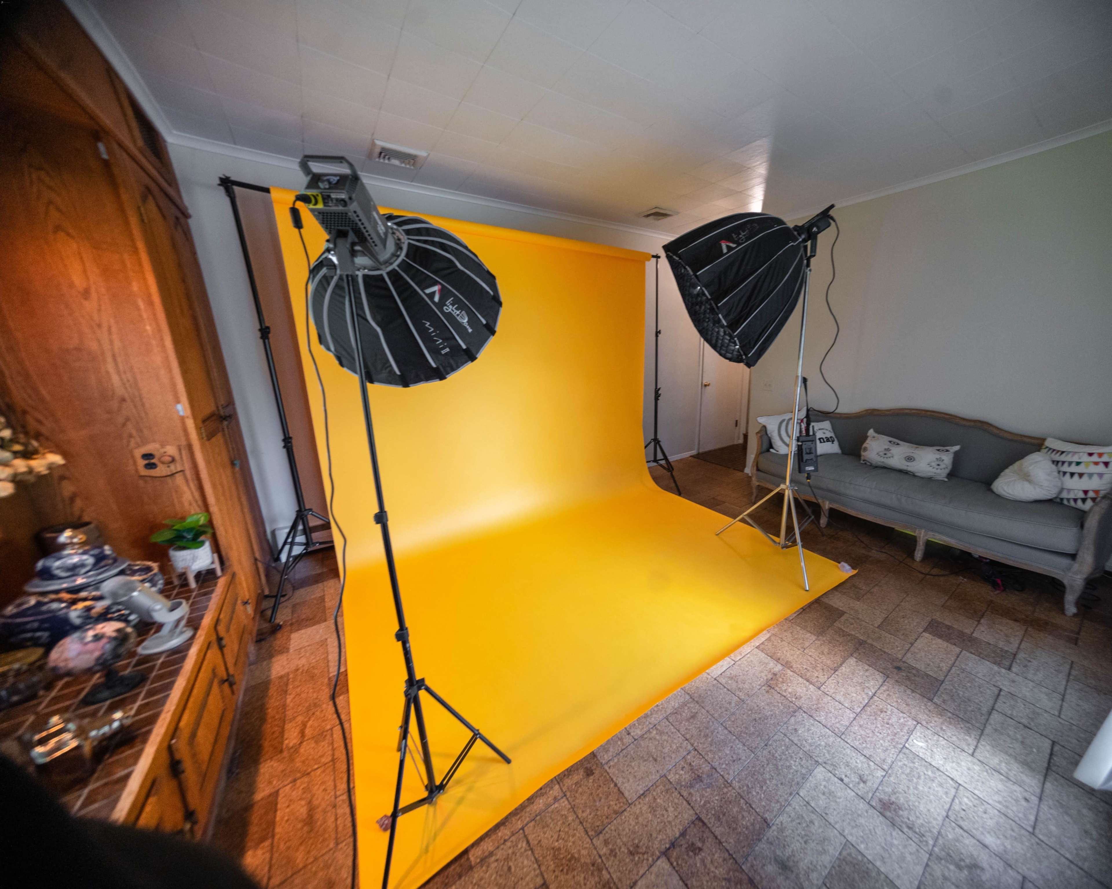 A photography setup featuring two studio lights positioned toward a yellow backdrop in a room with a wooden cabinet and a couch.