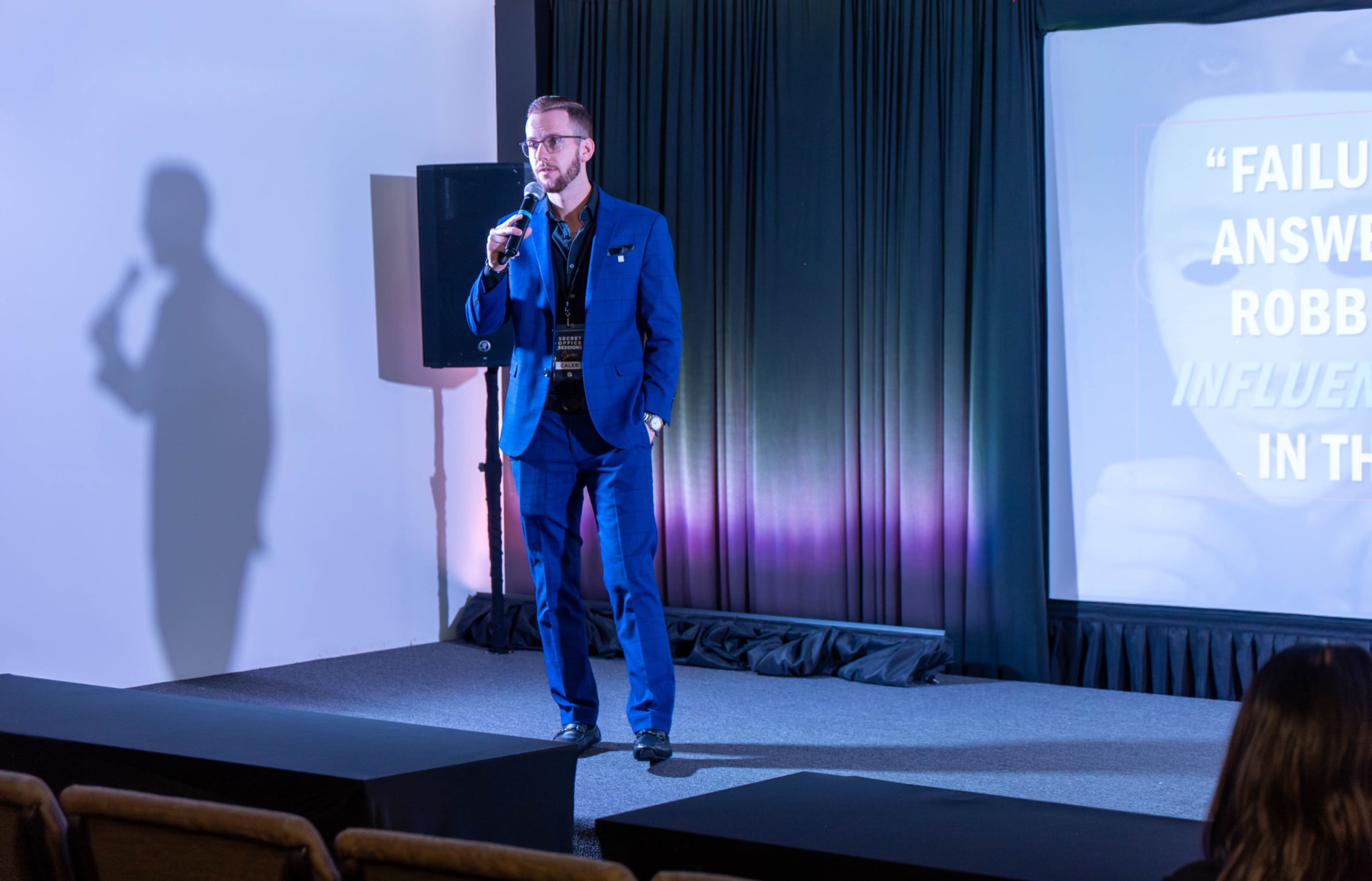 A man in a blue suit is speaking into a microphone on stage, with a presentation screen and lighting in the background.