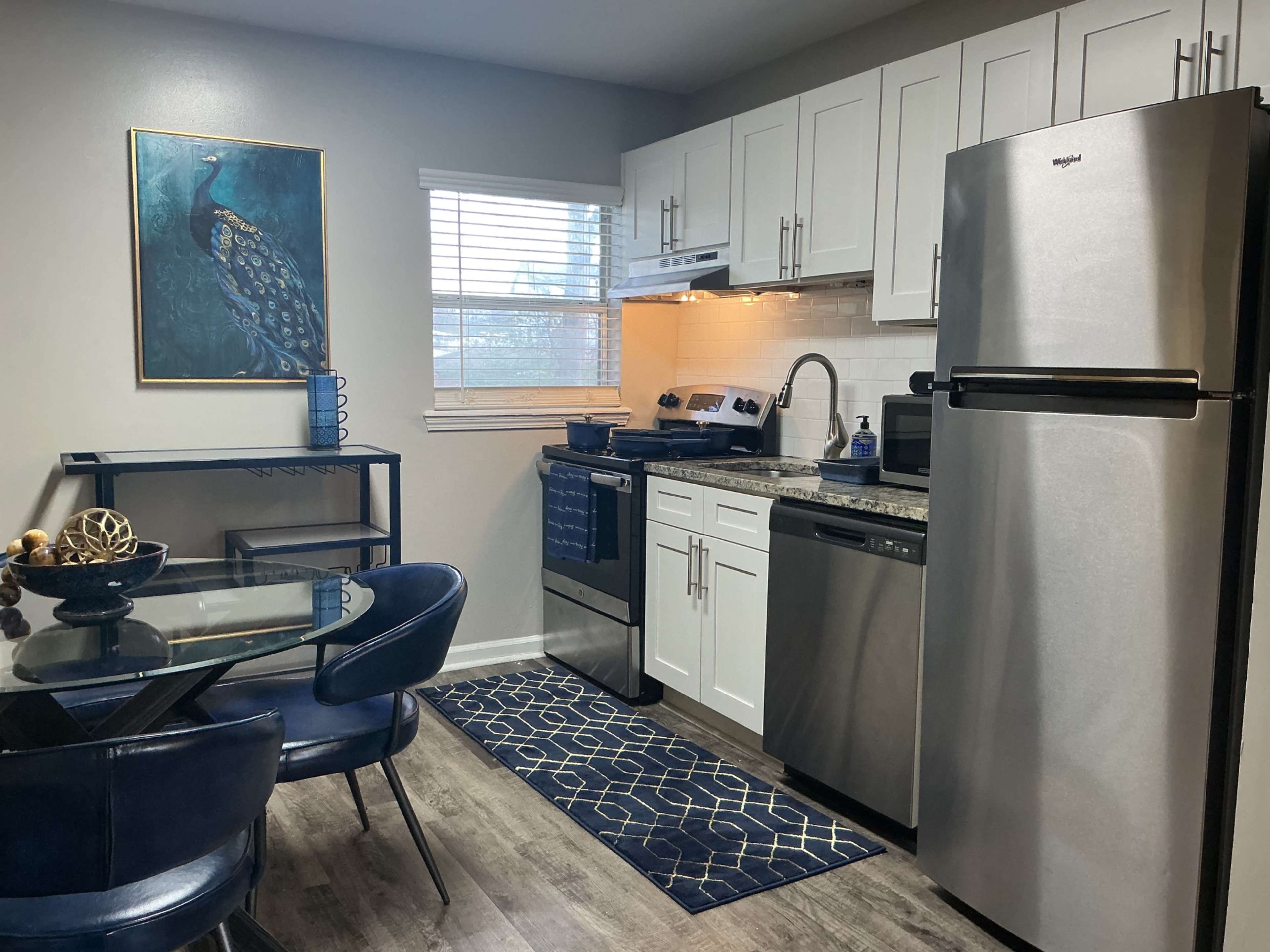 The image shows a modern kitchen with stainless steel appliances, white cabinets, a dining table with blue chairs, and a peacock wall art.