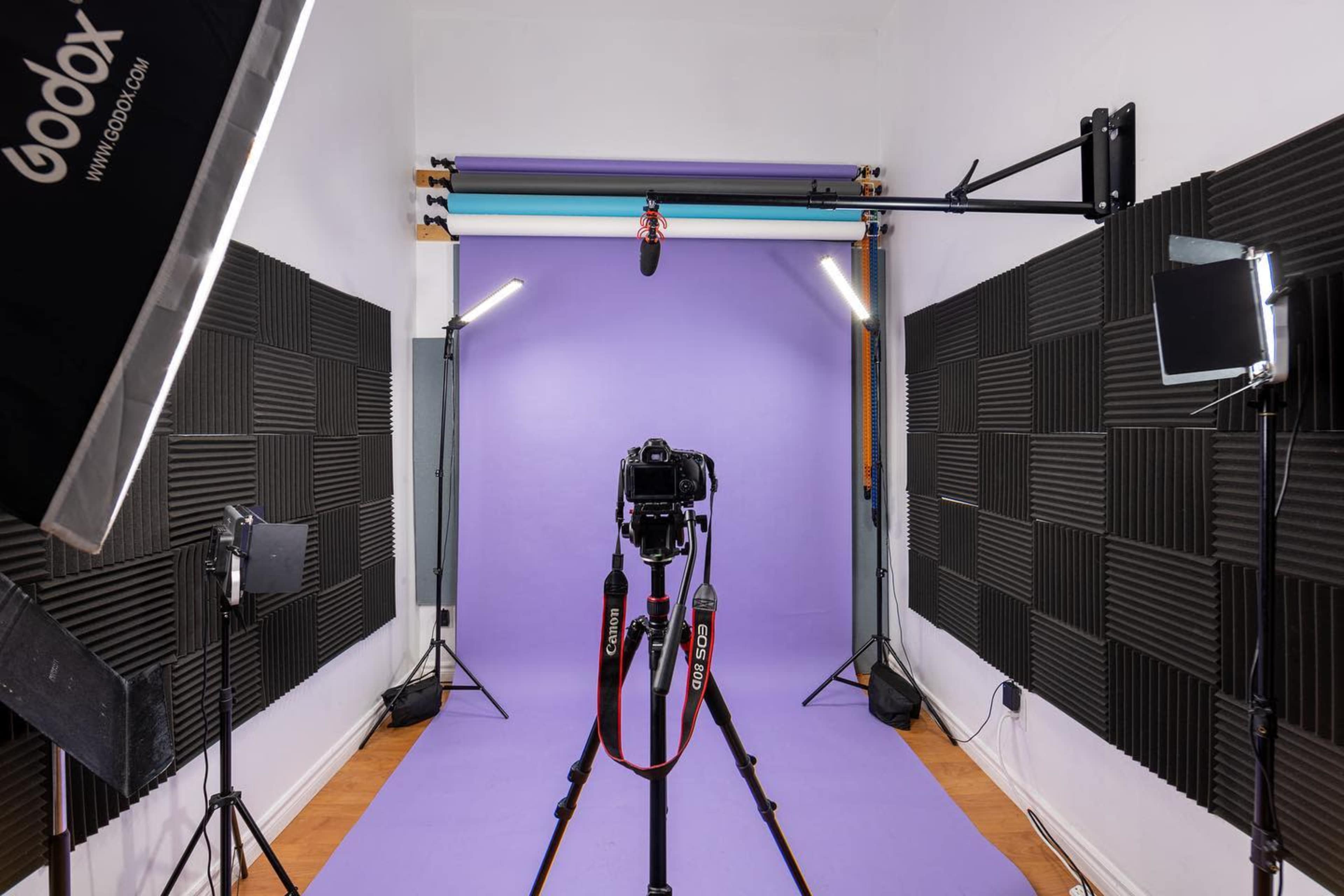 A photography studio features a camera on a tripod facing a purple backdrop, surrounded by various lighting equipment.
