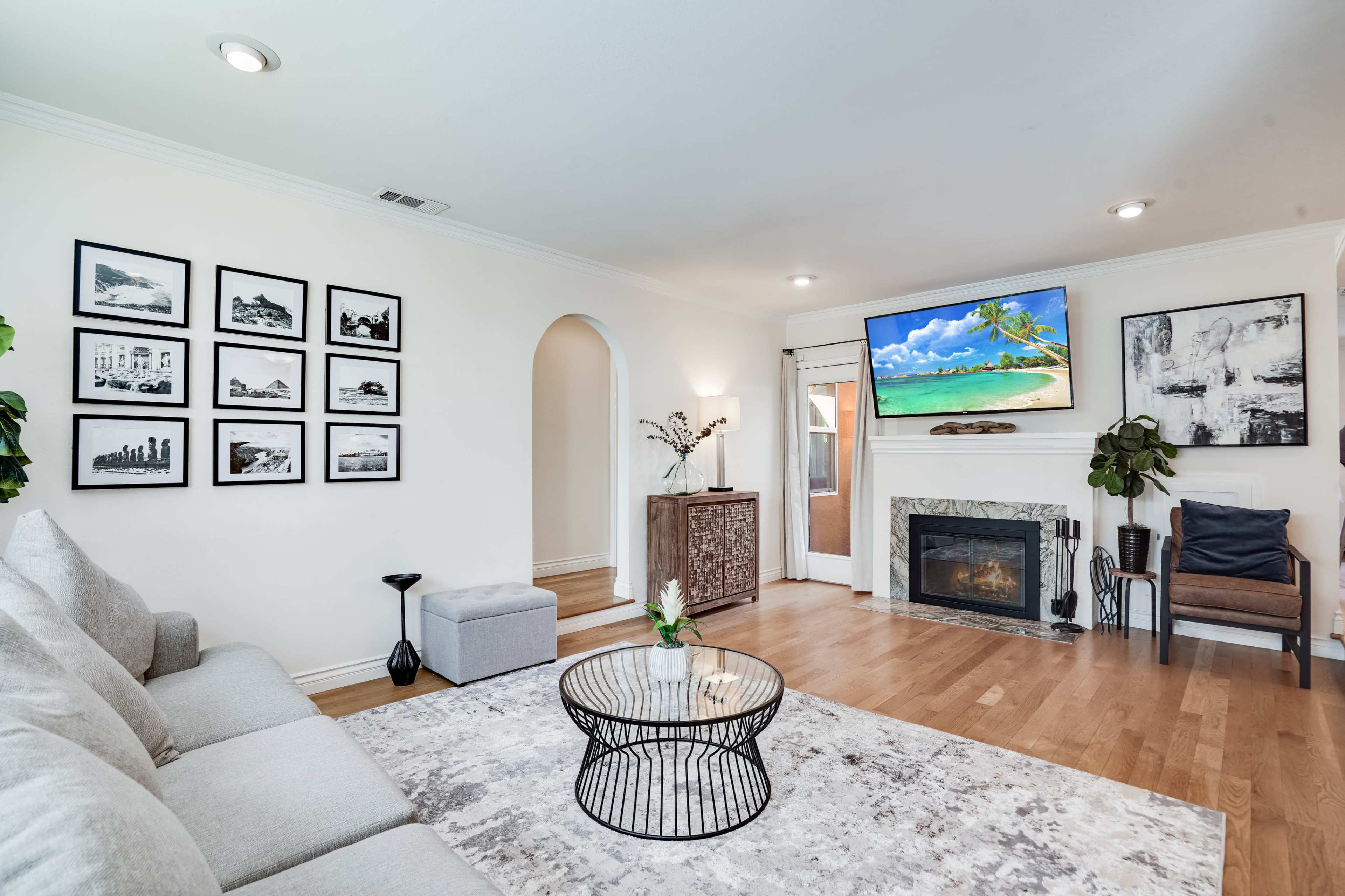 A living room features a gray sectional sofa, a round coffee table, a mounted television displaying a beach scene, and a fireplace with surrounding decorative elements, all illuminated by natural light.