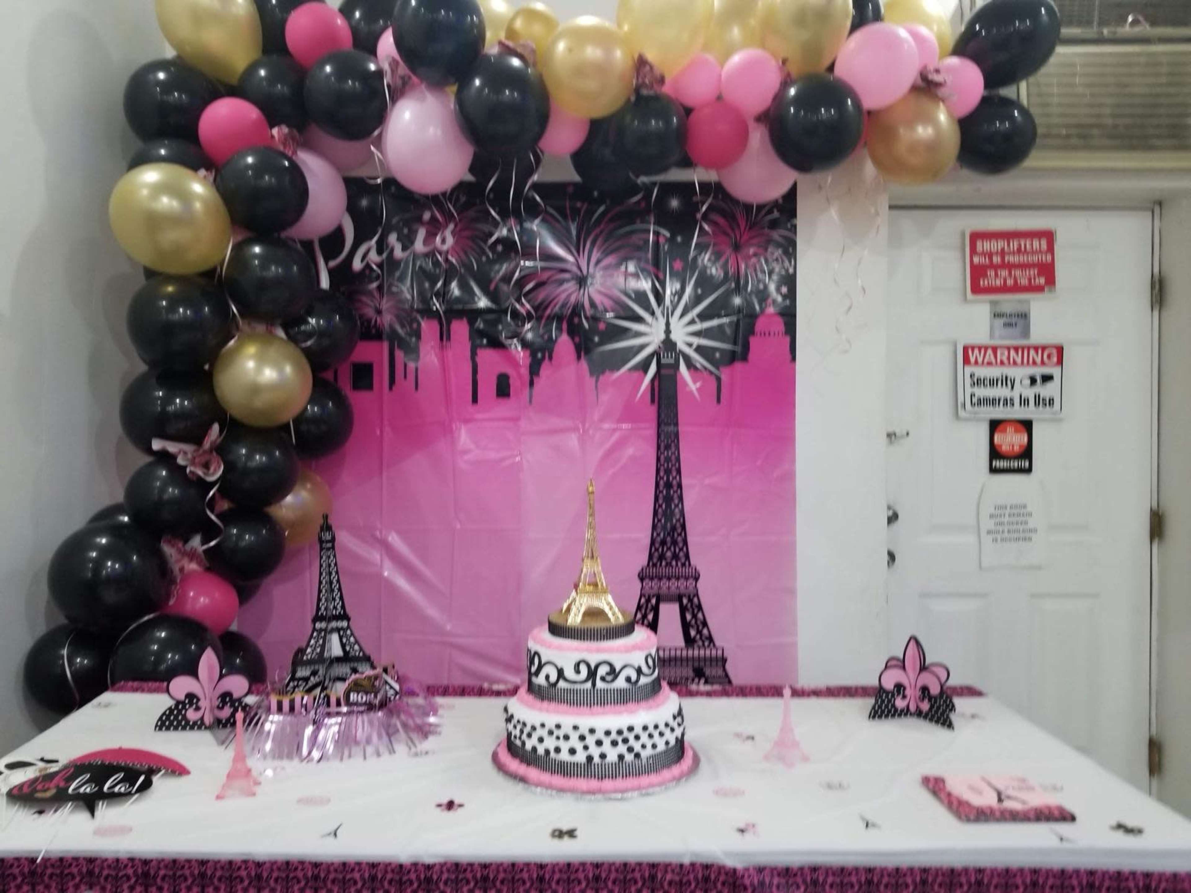 A decorated table displays a cake featuring an Eiffel Tower design, surrounded by balloons and a backdrop themed with Parisian landmarks.