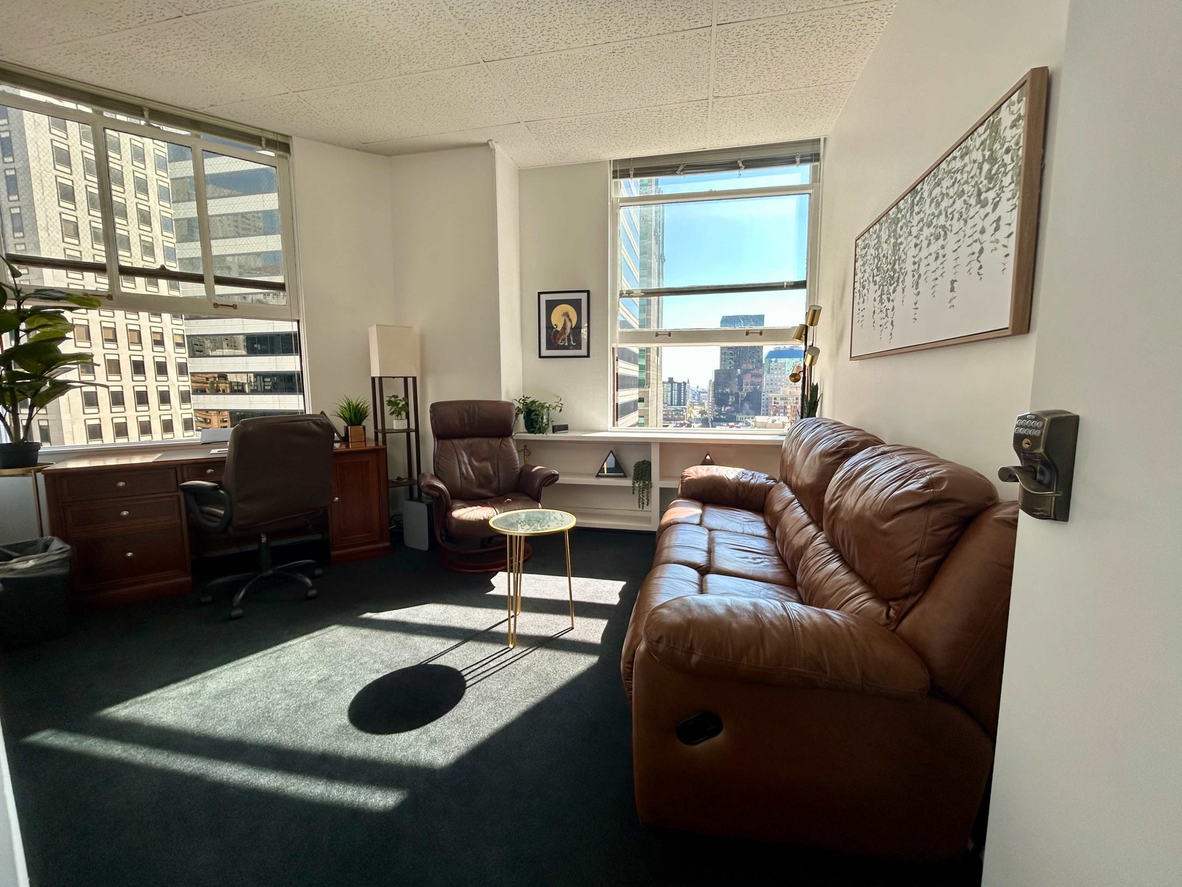 A modern office space with a brown leather sofa, a glass coffee table, and a desk, bathed in natural light from large windows.