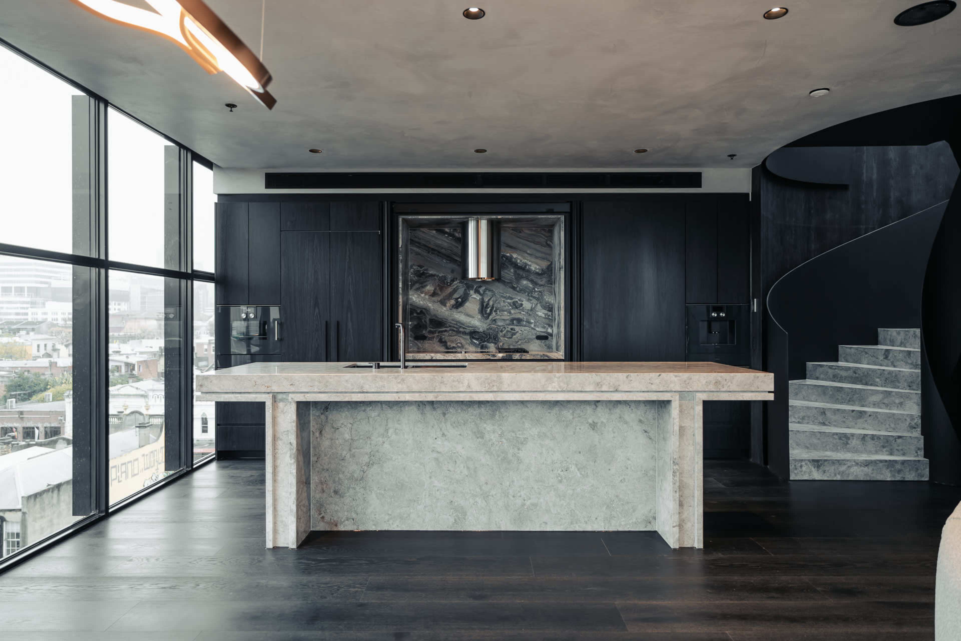 A modern kitchen features a large stone island, dark cabinetry, and a spiral staircase in the background.