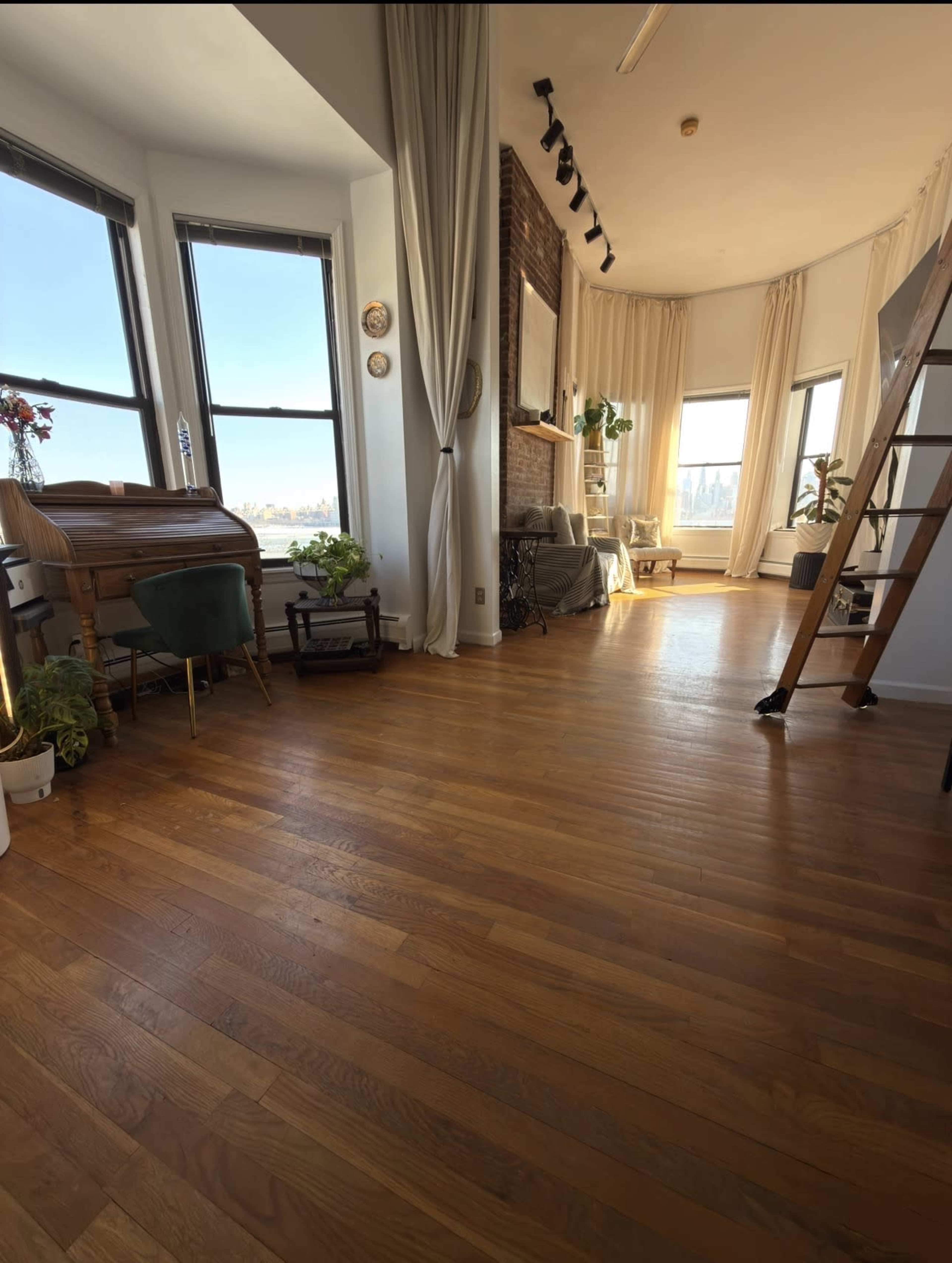 The image shows a spacious room with large windows, wooden flooring, and a cozy arrangement of furniture, including a piano and houseplants.