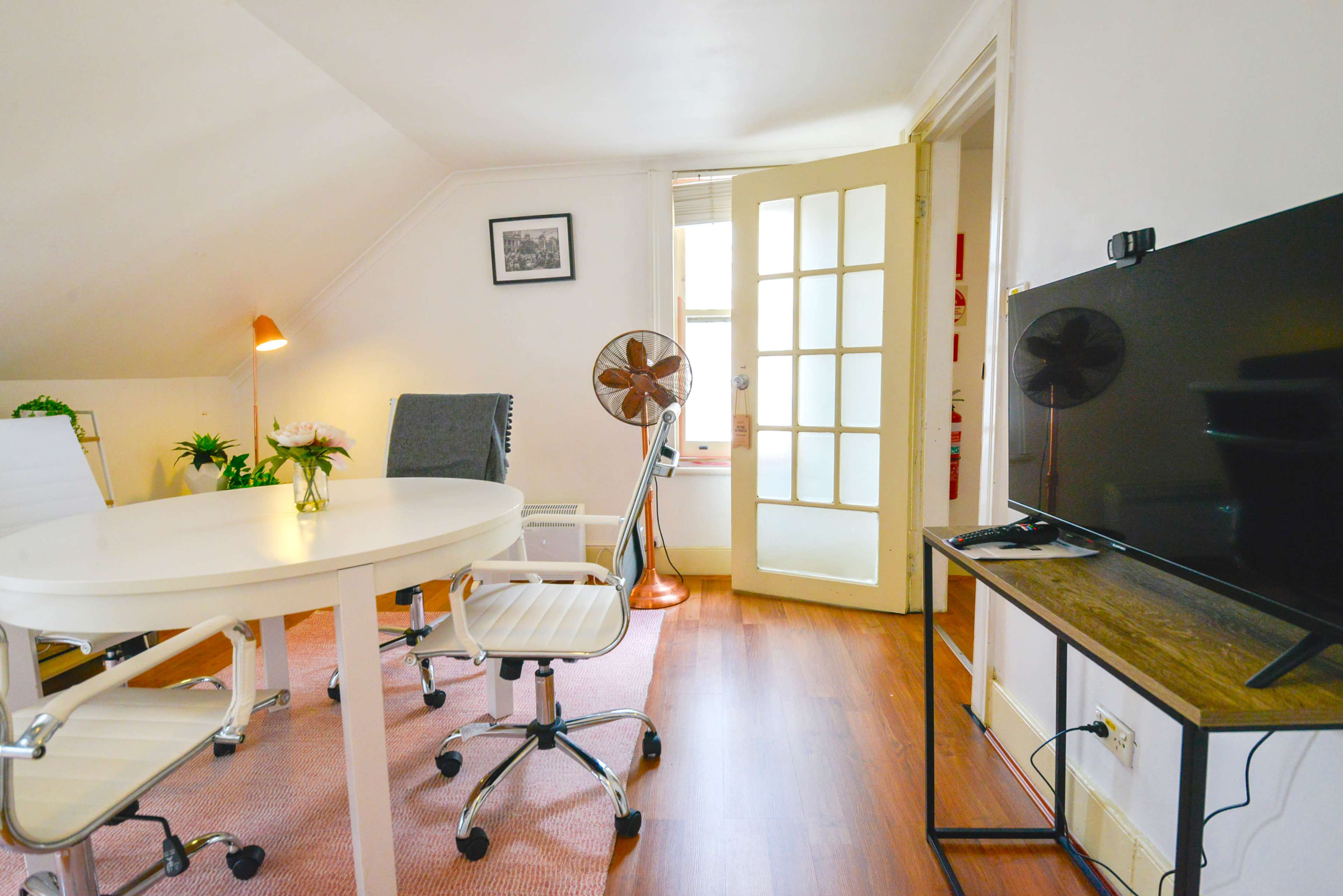 A small office space with a round table, two white chairs, and a television mounted on a wooden stand, illuminated by natural light through a door leading outside.