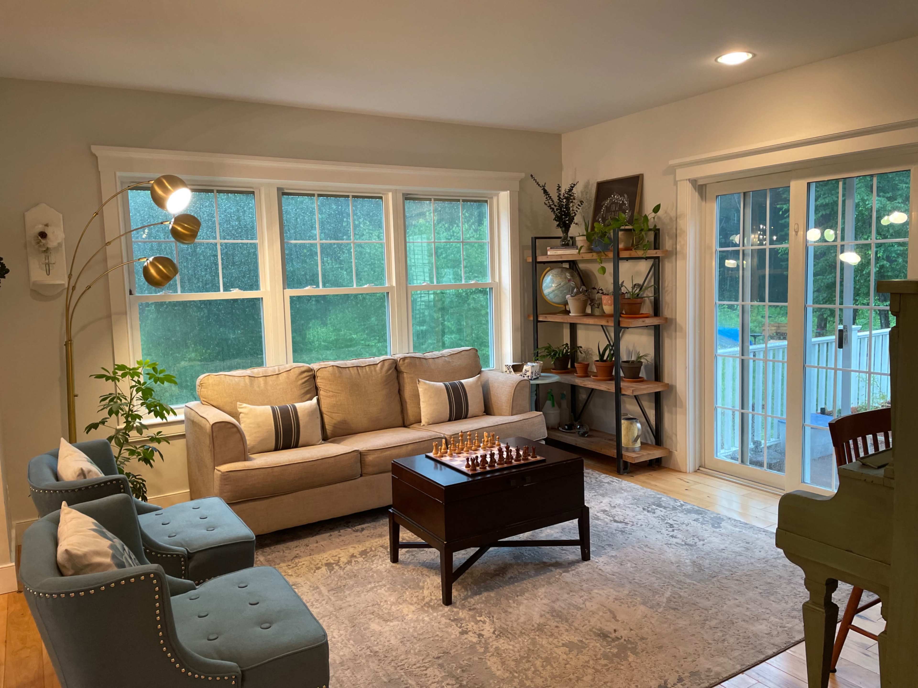 The image shows a cozy living room with large windows, a beige sofa, a wooden coffee table with a chess set, blue armchairs, and shelves displaying decorative items.