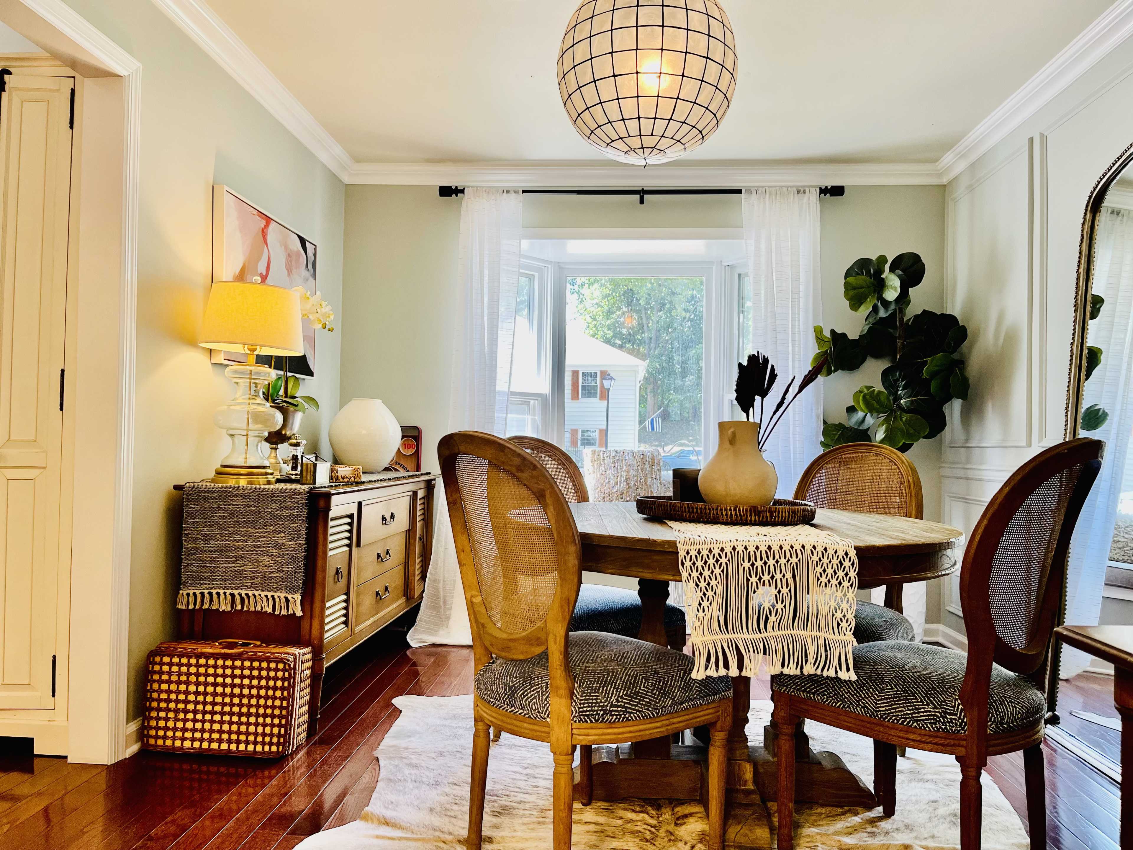 A round wooden dining table with woven chairs is surrounded by a light-filled room featuring a large window and decorative plants.