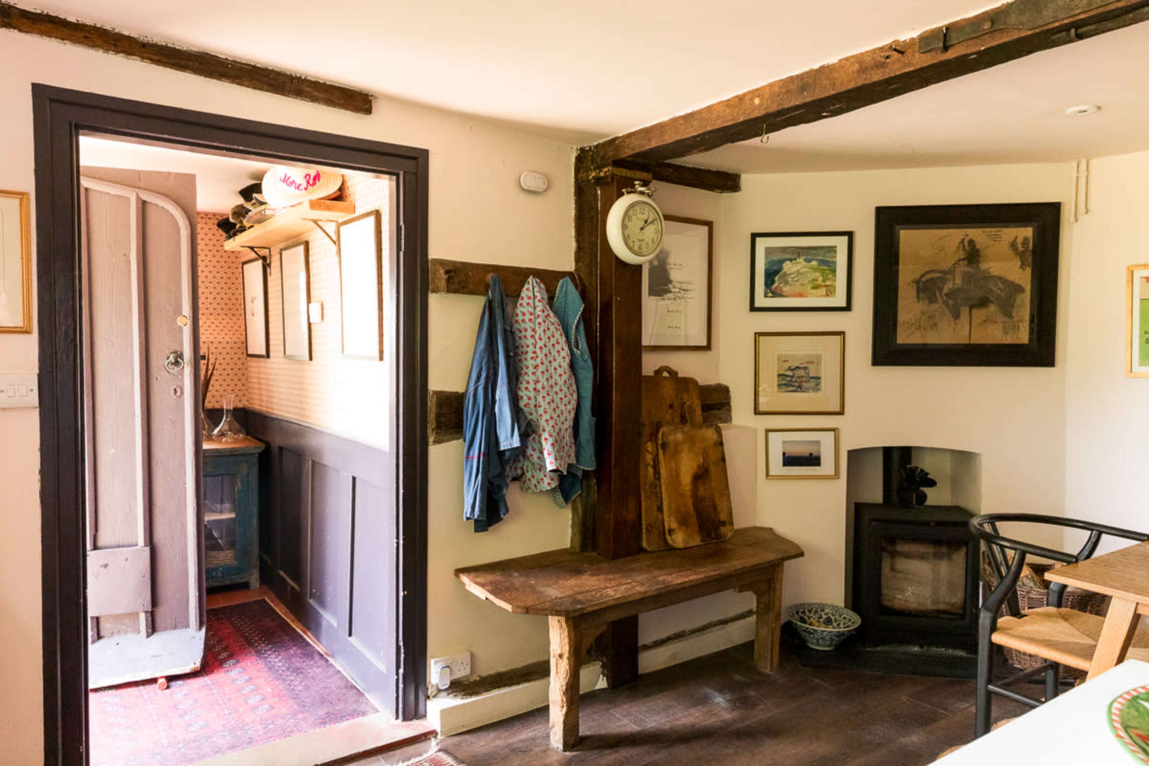 The image shows a cozy interior of a room with a wooden bench, a small stove, and various framed artworks on the walls, alongside a doorway leading to another room.