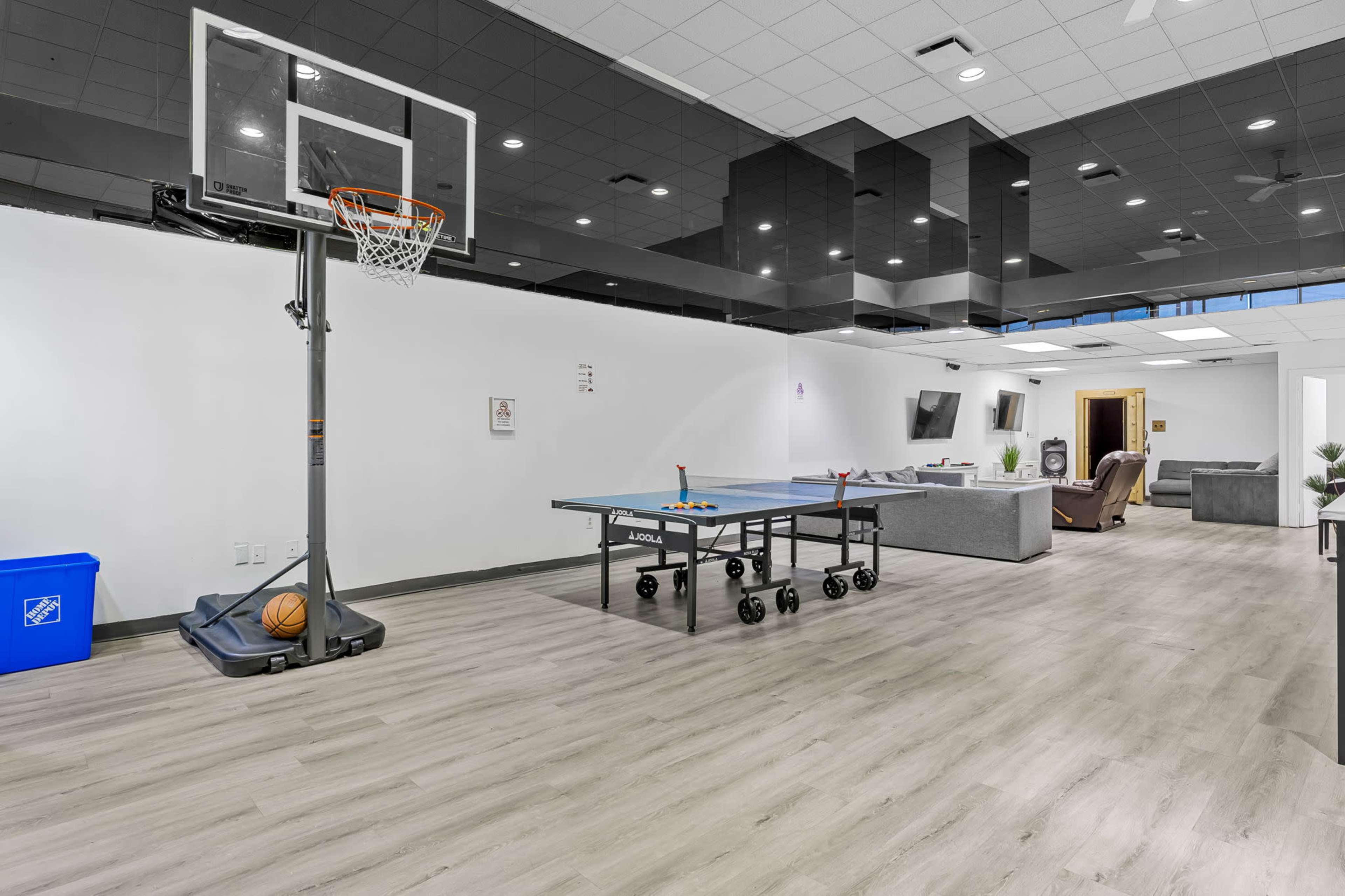 The image shows a recreational room featuring a basketball hoop, a ping pong table, and comfortable seating along with a large flat-screen TV.