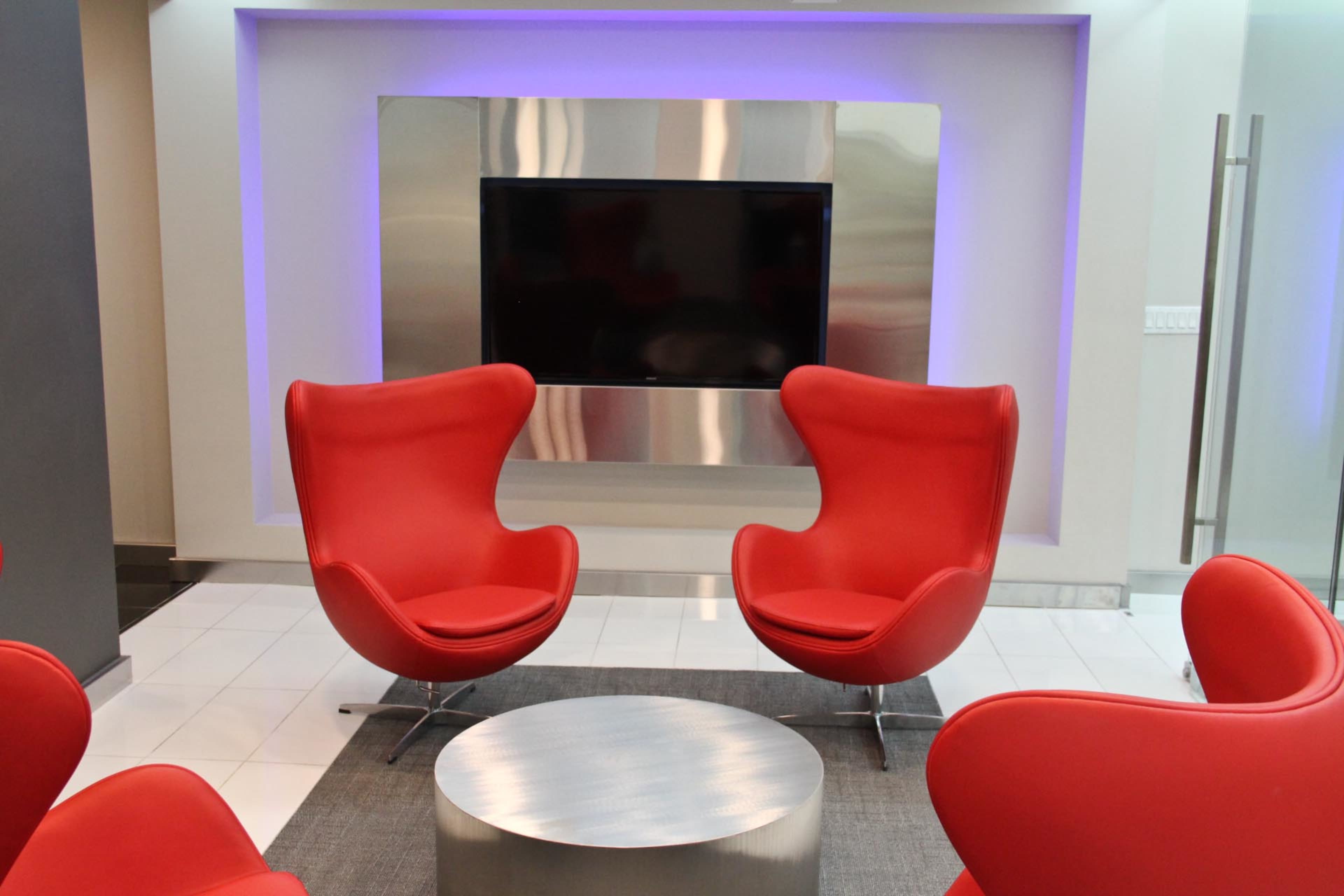 The image shows a modern lounge area featuring two red chairs and a circular silver table in front of a flat-screen TV mounted on a sleek wall.