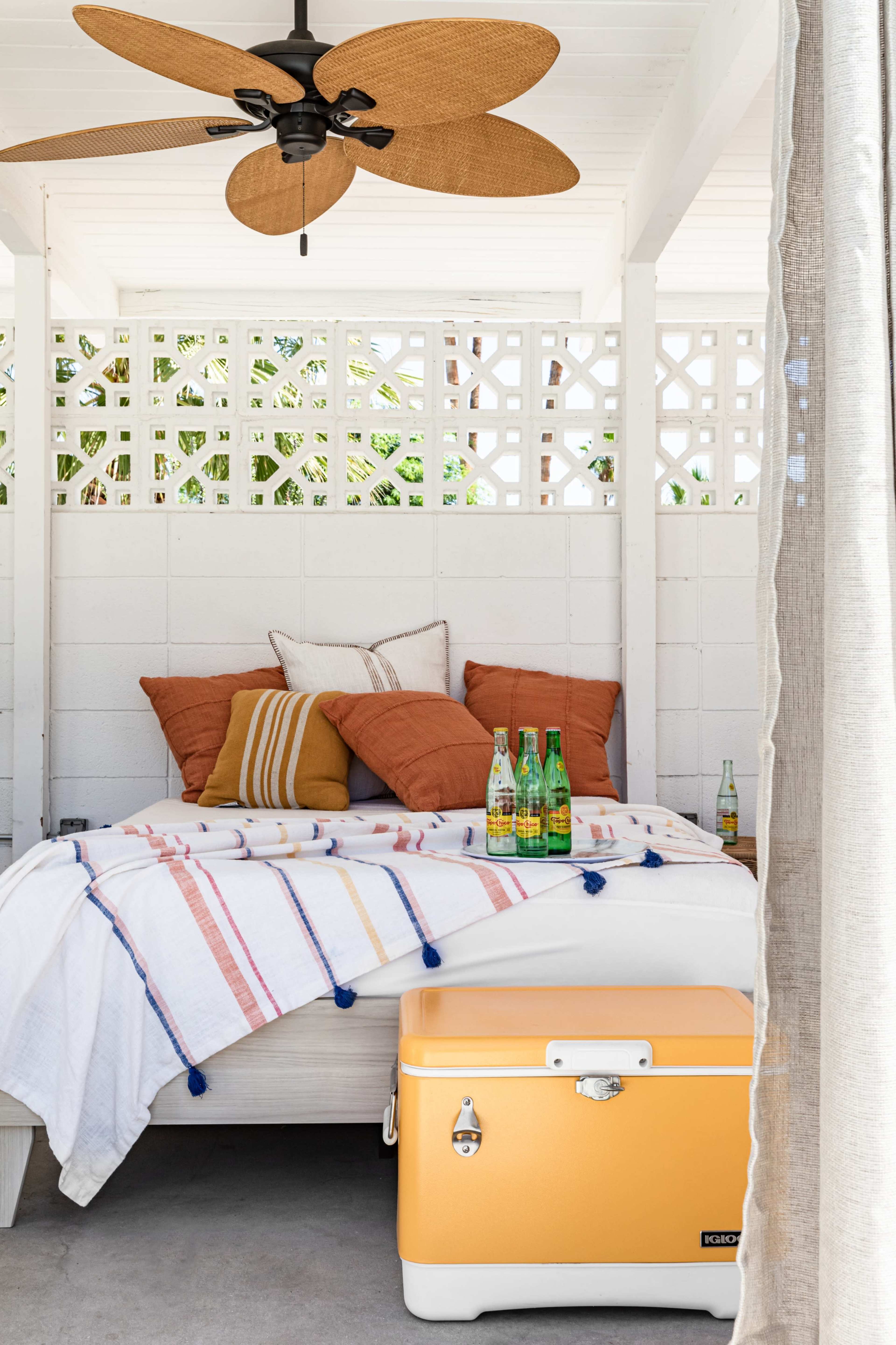 The image shows a cozy outdoor space featuring a bed with orange and white pillows, a colorful blanket, and a yellow cooler with drinks.