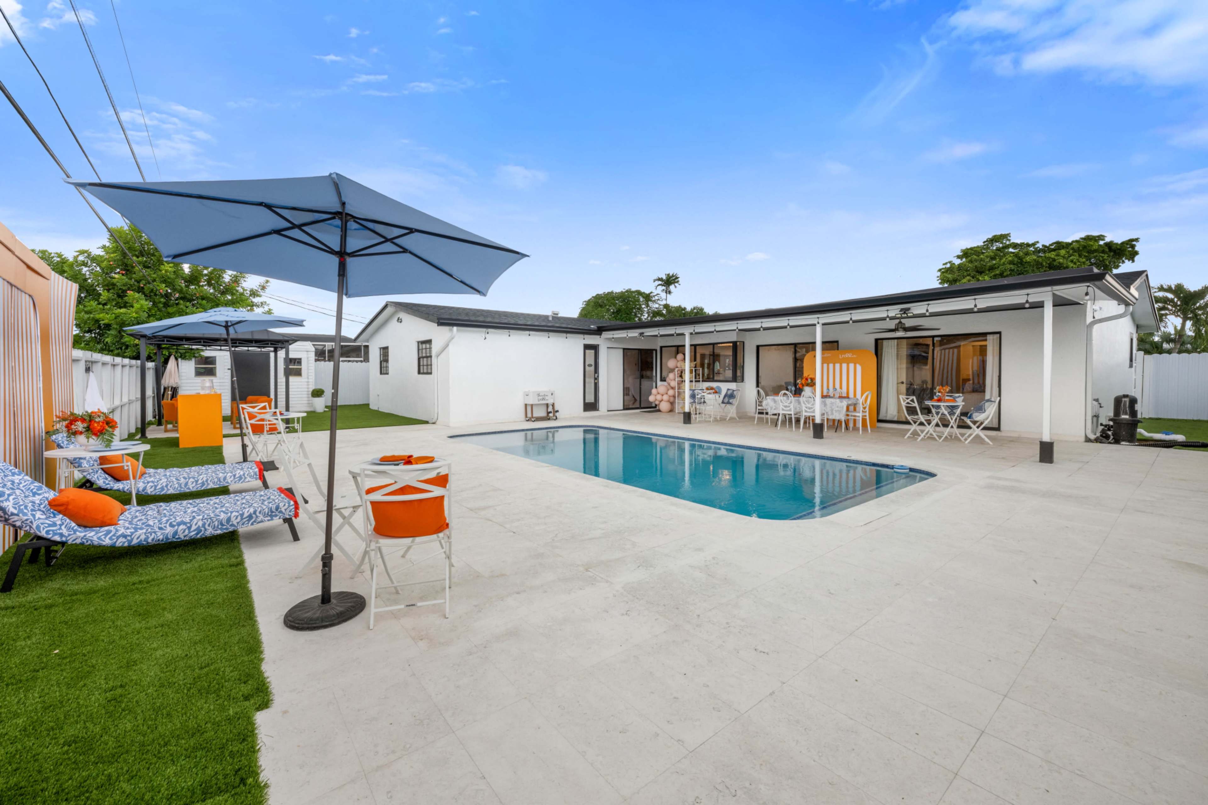 A modern backyard features a swimming pool surrounded by lounge chairs, patio tables, and umbrellas, with a sleek house in the background.